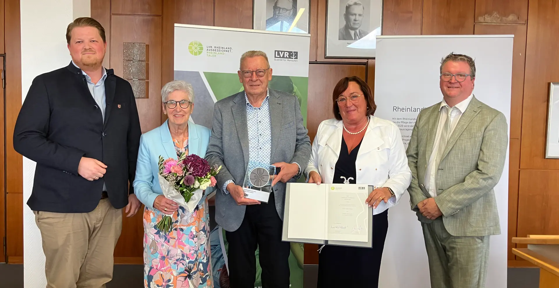 Ein Gruppenfoto von zwei Frauen und drei Männer. Die eine Frau hält einen Blumenstrauß in der Hand, die andere eine Urkunde. Der Mann in der Mitte hält eine Trophäe.