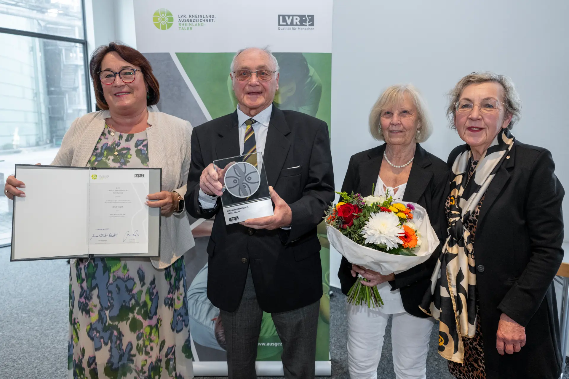 Drei Frauen und ein Mann stehen vor einem Aufsteller mit dem Rheinland ausgezeichnet-Logo. Die Frau links hält eine Urkunde in der Hand, der Mann rechts daneben eine Trophäe und die Frau daneben einen Blumenstrauß.