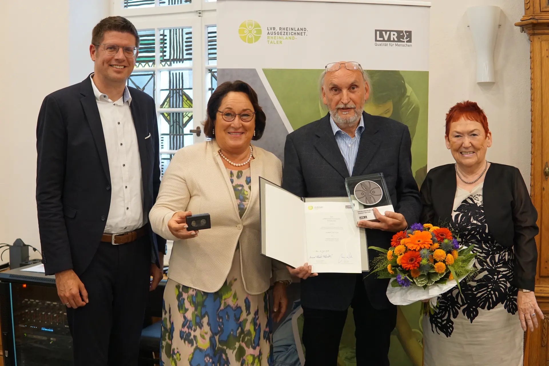 Zwei Frauen und zwei Männer. Die Frau ganz rechts hält einen Blumenstrauß. Der Mann links daneben hält eine Urkunde und eine Trophäe.