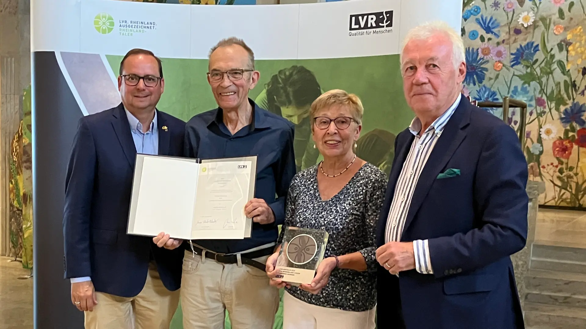 Drei Männer und eine Frau lächeln in die Kamera. Der Mann in der Mitte hält eine Urkunde in der Hand, die Frau rechts daneben eine Trophäe.