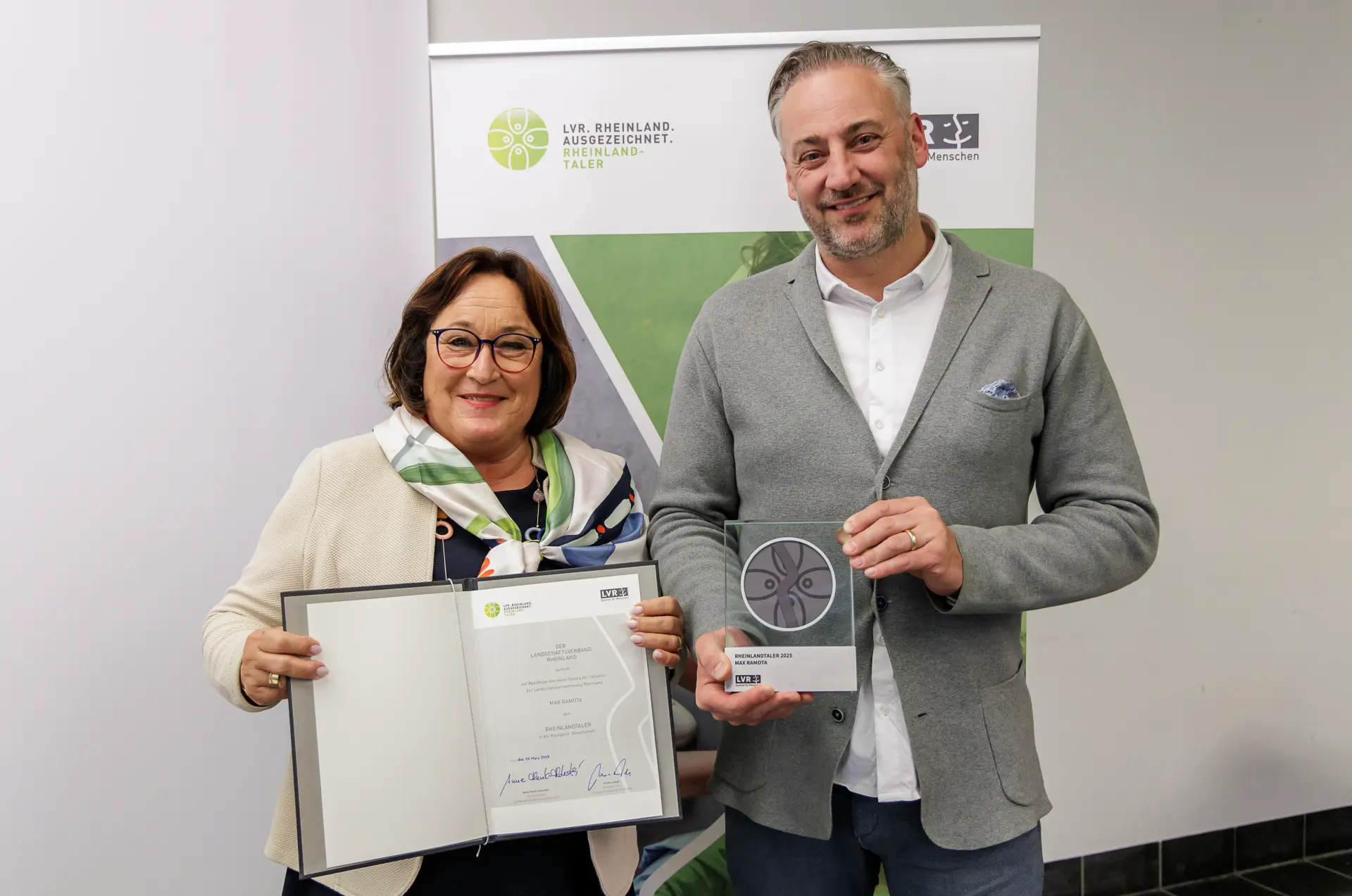 Eine Frau hält eine Urkunde in der Hand. Der Mann rechts daneben eine Trophäe. Hinter den beiden steht ein Aufsteller mit dem Rheinland ausgezeichnet-Logo.