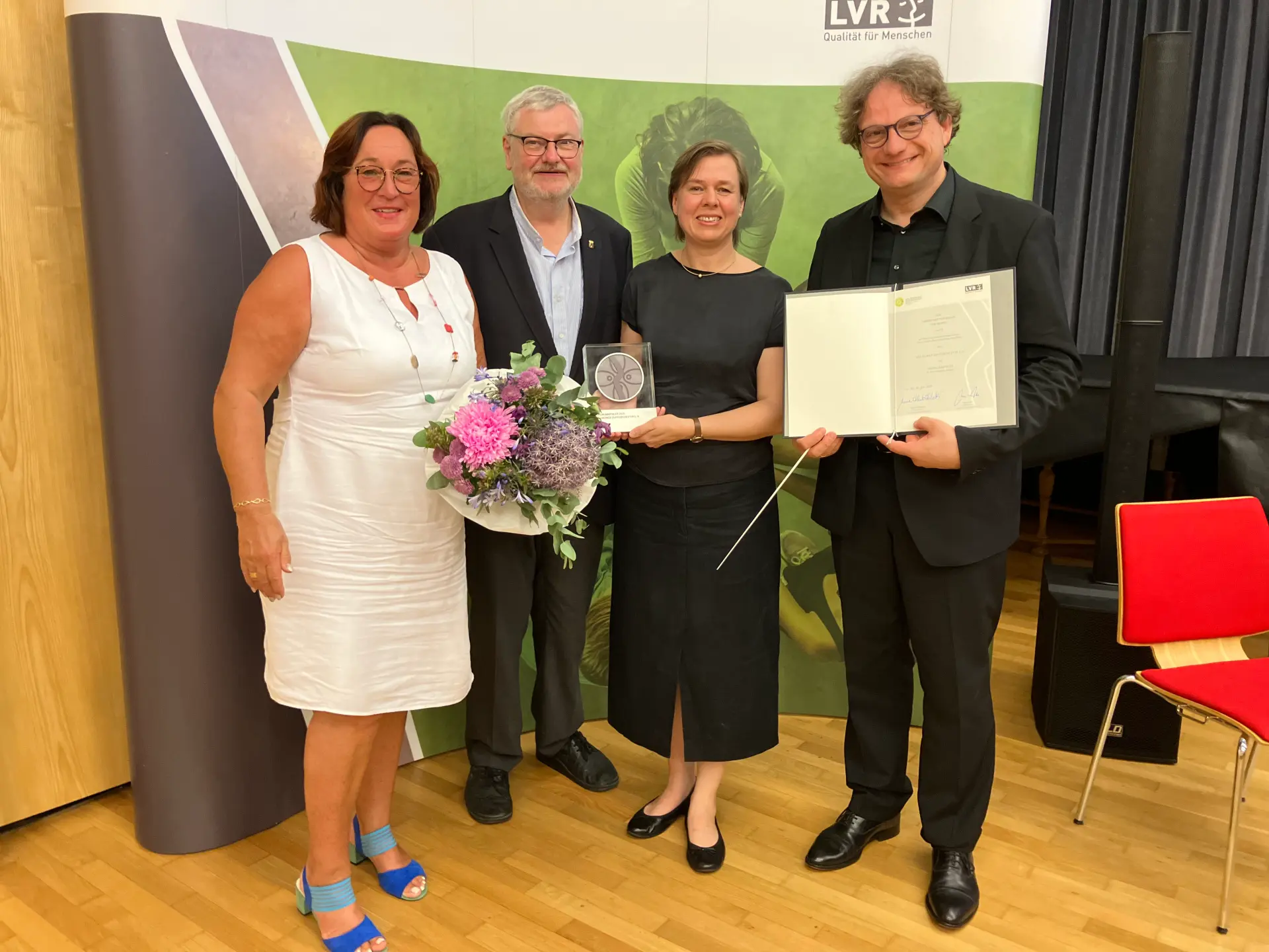 Zwei Frauen und zwei Männer stehen vor einer Leinwand. Der Mann rechts hält eine Urkunde, die Frau links daneben eine Trophäe.