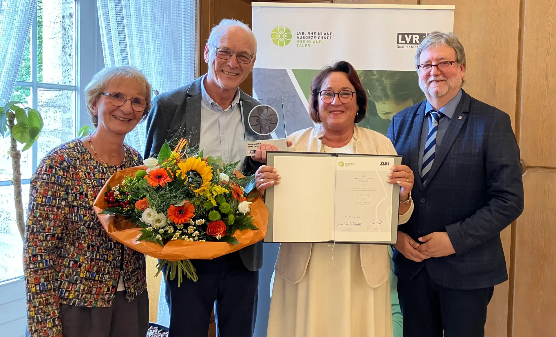 Zwei Frauen und zwei Männer lächeln in die Kamera. Eine Frau hält einen Blumenstrauß, die andere eine Urkunde.