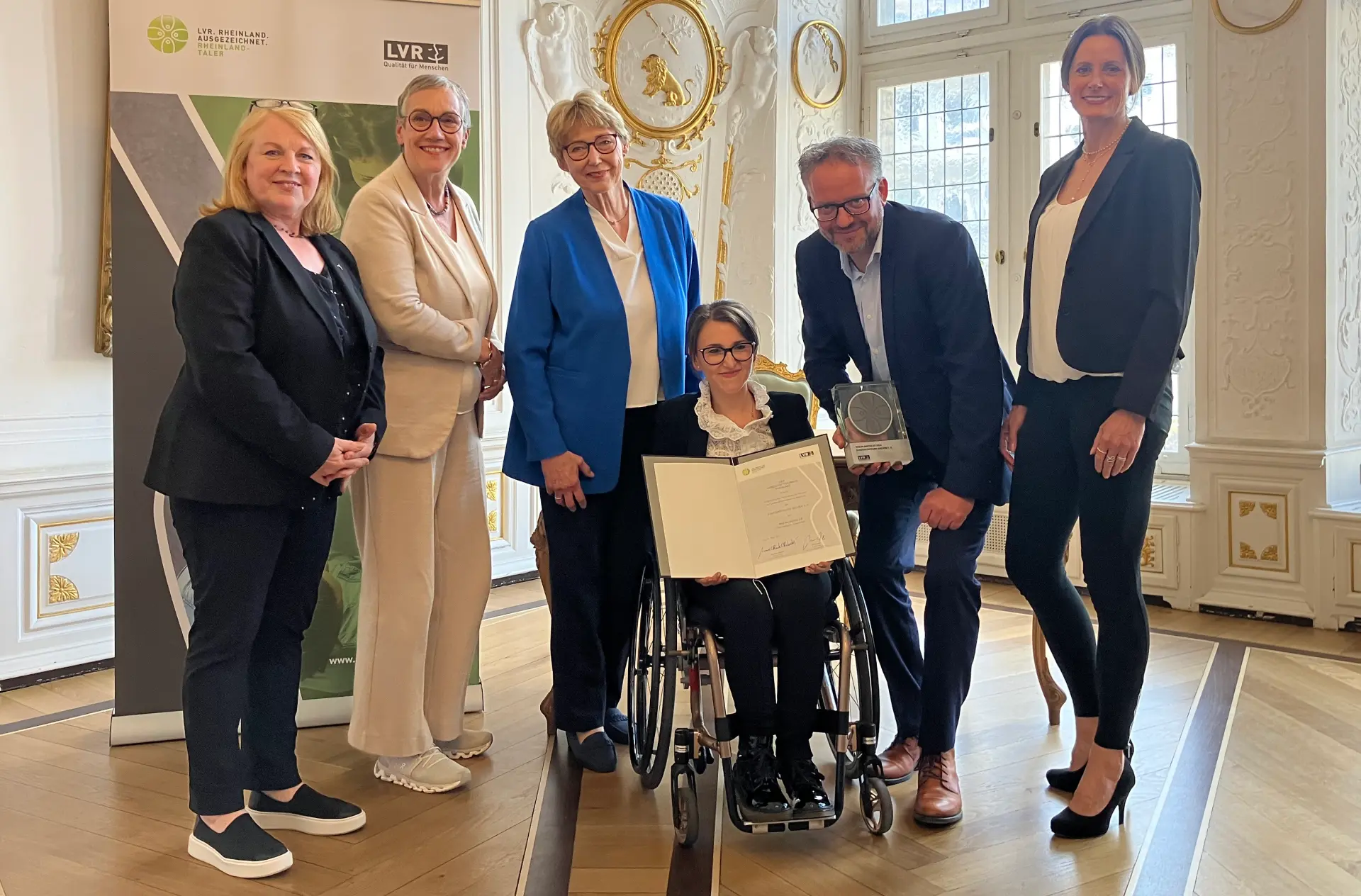 Ein Gruppenfoto. Die Frau in der Mitte sitzt im Rollstuhl. Sie hält eine Urkunde in der Hand.
