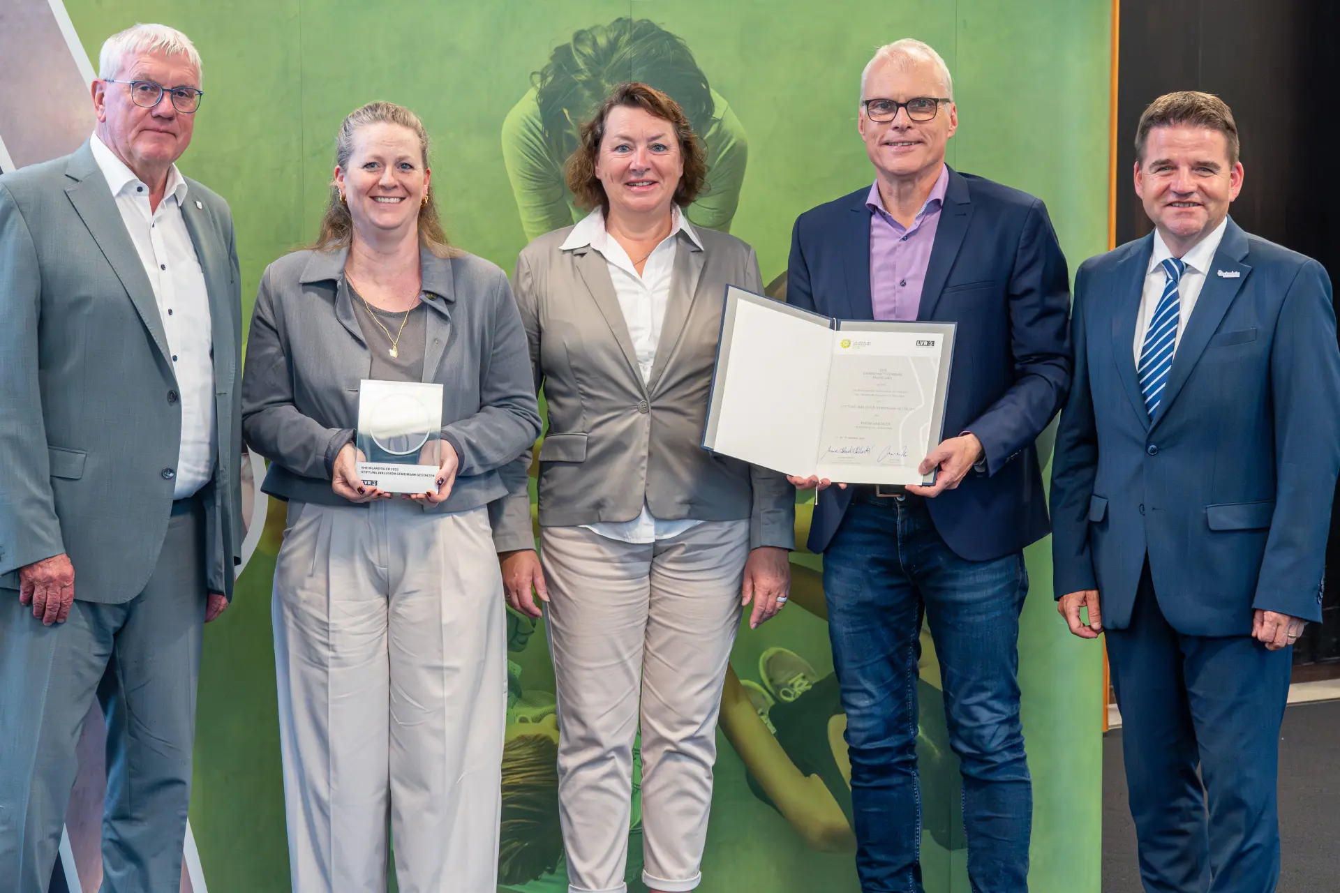 Zwei Frauen und drei Männer stehen vor einer grünen Leinwand. Eine Frau hält eine Trophäe, ein Mann hält eine Urkunde in der Hand.