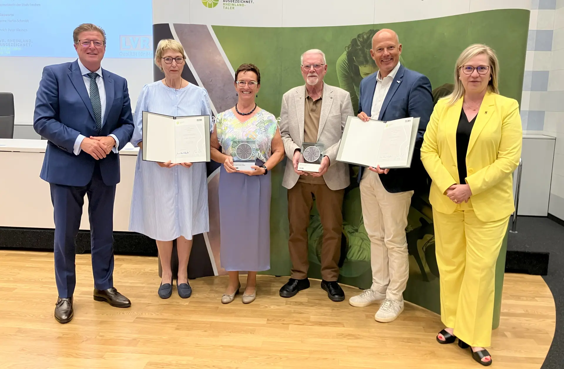 Ein Gruppenfoto vor dem Rheinland ausgezeichnet-Aufsteller. Die Frau und der Mann in der Mitte halten jeweils eine Trophäe in der Hand.