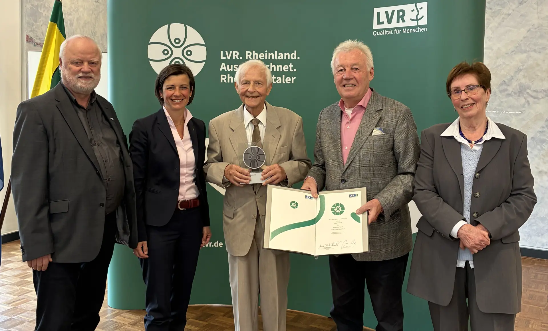 Eine Gruppe Menschen steht vor einem grünen Hintergrund mit dem Logo des Rheinlandtalers. Der Mann in der Mitte hält eine Trophäe in der Hand.
