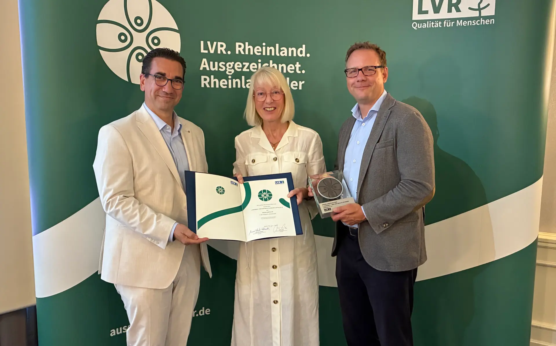 Zwei Männer und eine Frau vor einer grünen Leinwand. Der Mann links hält eine Trophäe in der Hand.