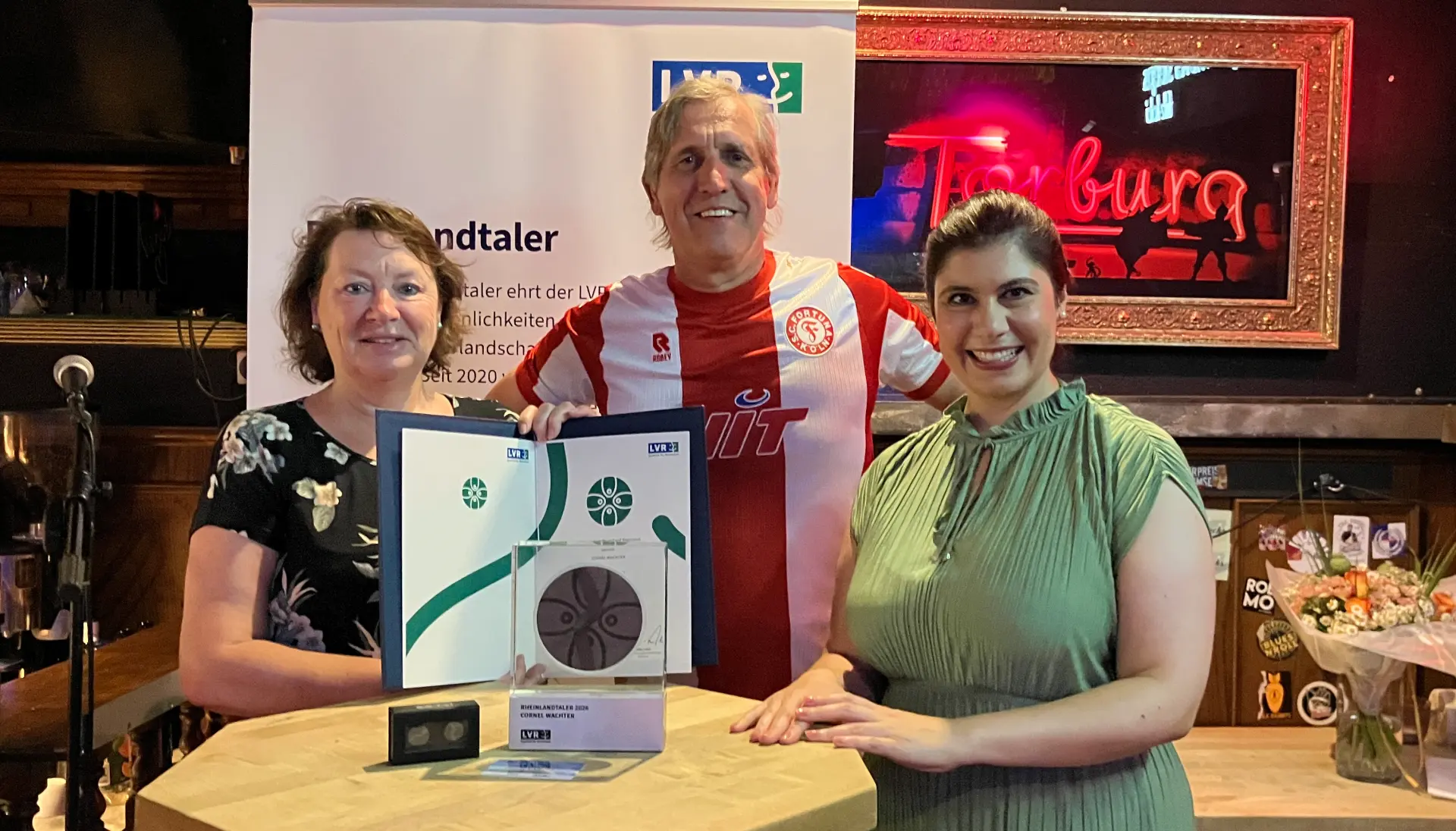 Zwei Frauen und ein Mann stehen an einem Stehtisch. Auf dem Tisch steht eine Trophäe. Die Frau ganz links hält eine Urkunde.