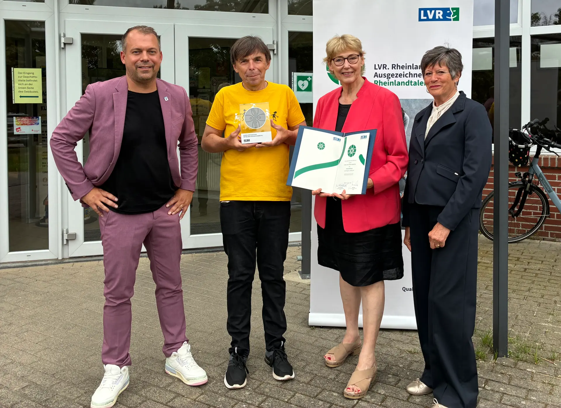 Eine Gruppe Menschen steht vor einem Vereinsheim. Der zweite Mann von links hält eine Trophäe in der Hand.