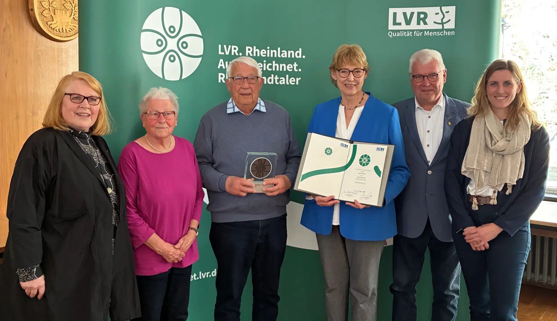 Drei Frauen und drei Männer stehend vor einer Wand; eine Frau hält Urkunde, ein Mann hält Trophäe