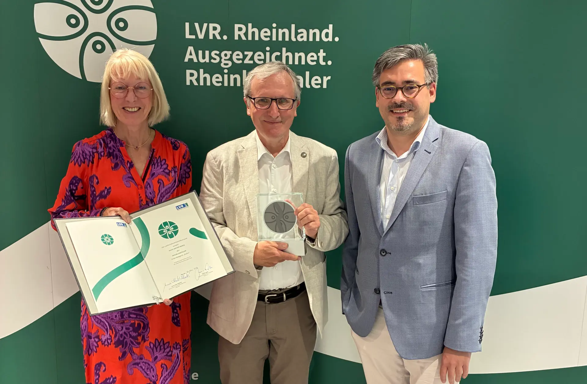 Eine Frau und zwei Männer stehen vor einer grünen Leinwand mit dem Rheinland ausgezeichnet-Logo. Der Mann in der Mitte hält eine Trophäe in der Hand. Die Frau links daneben eine Urkunde.