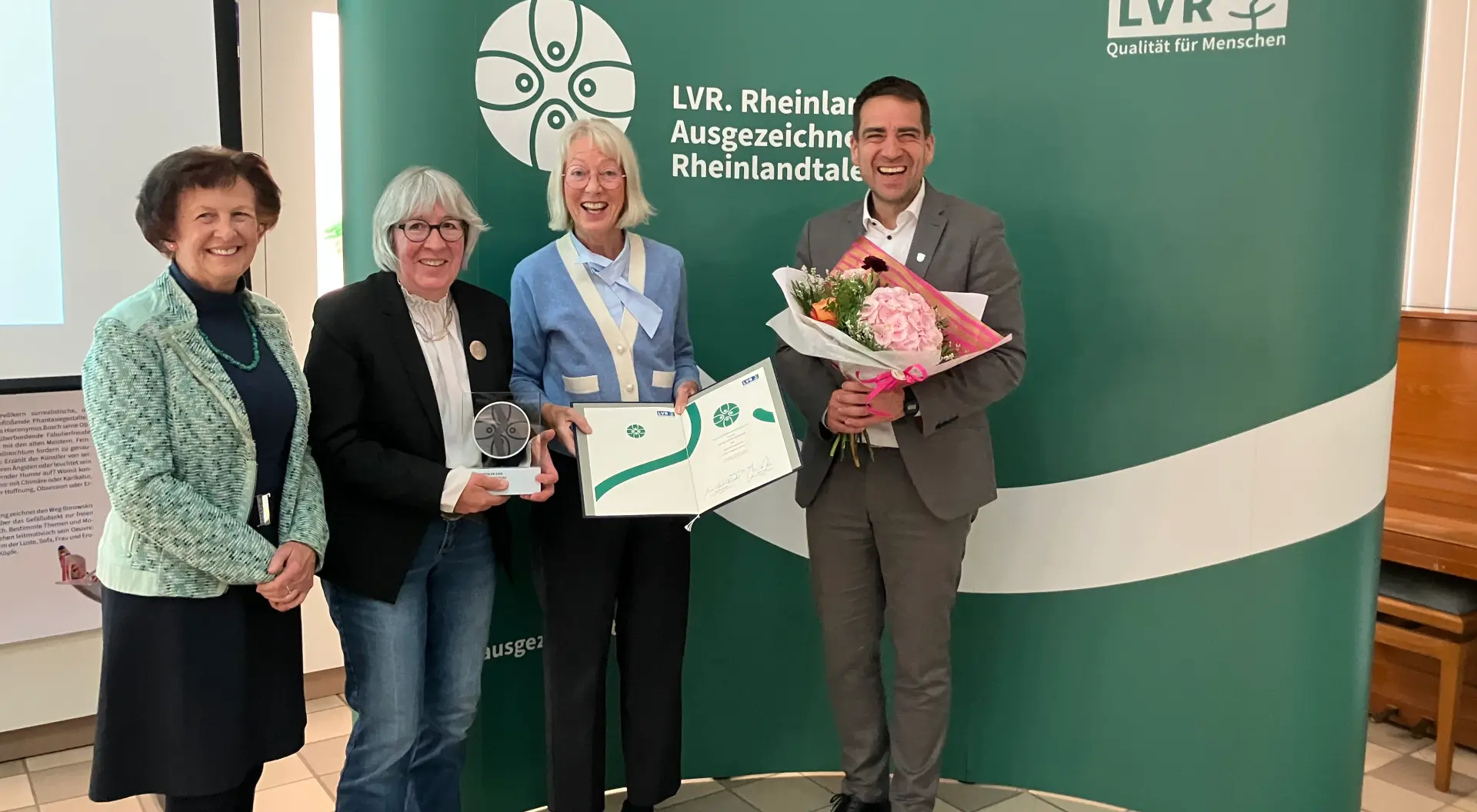 Drei Frauen und ein Mann stehen vor einem grünen Hintergrund mit dem Logo und dem Schriftzug des Rheinlandtalers. Die zweite Frau von links hält eine Trophäe in der Hand.