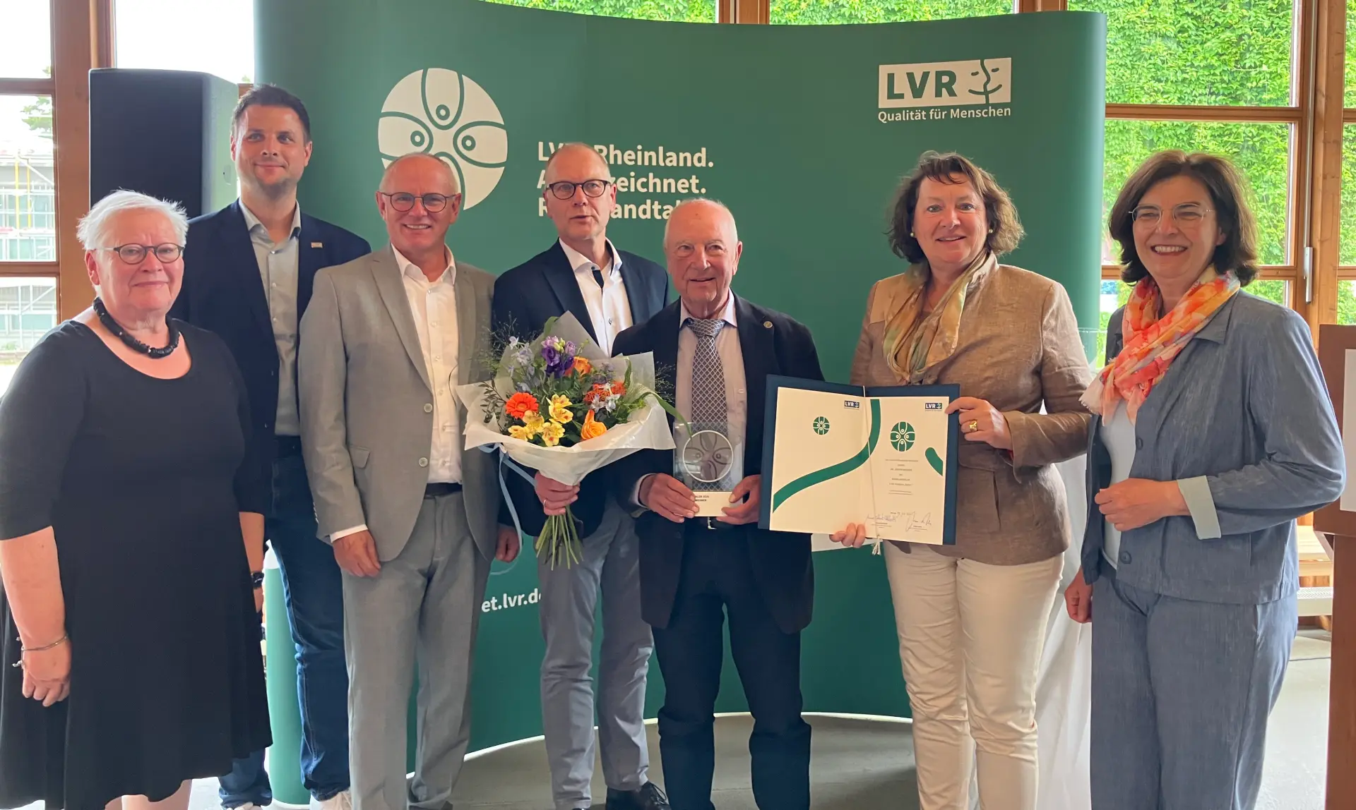 Ein Gruppenfoto vor einer grünen Leinwand. Der Mann in der Mitte hält eine Trophäe in der Hand. Der Mann links daneben einen Blumenstrauß. Die Frau rechts daneben eine Urkunde.