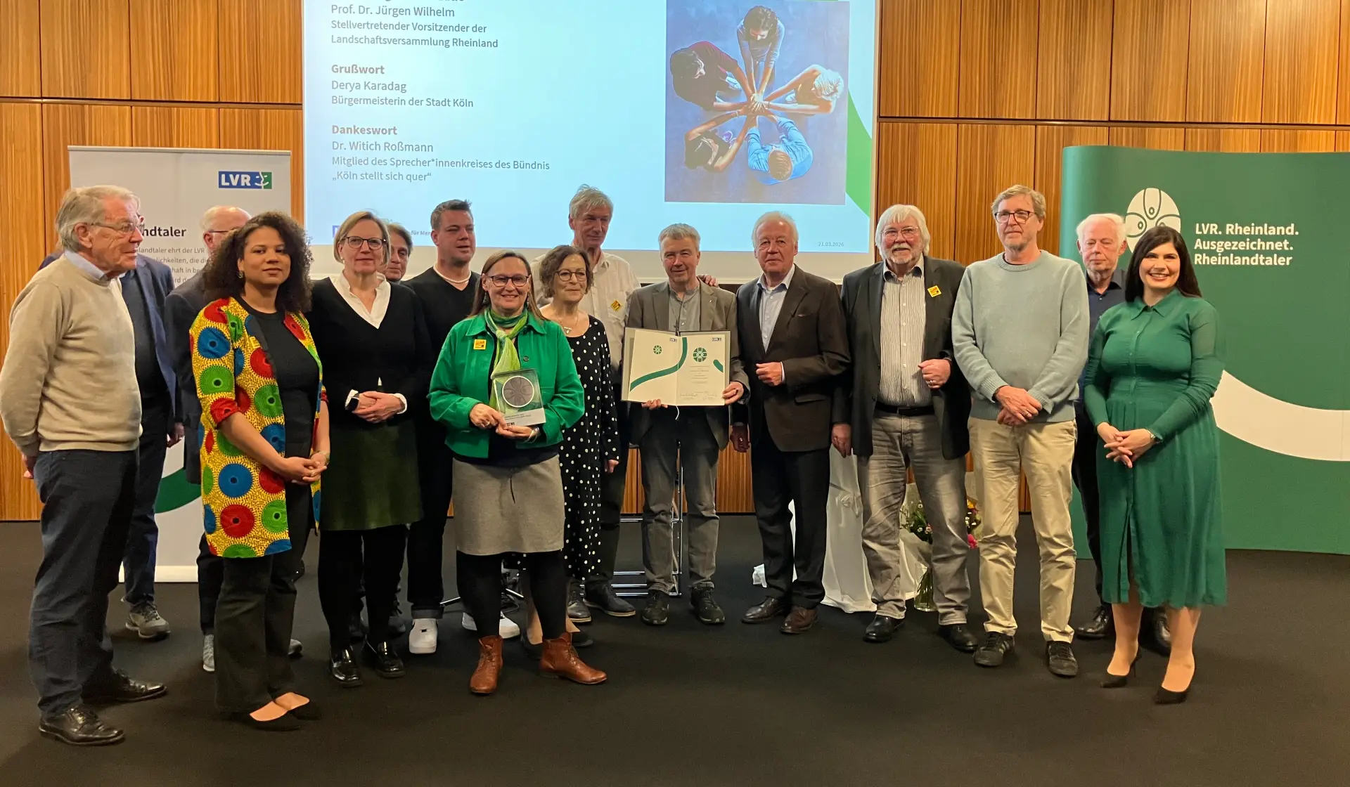Eine Gruppe Menschen, die vor einer Leinwand stehen. Eine Frau in der Mitte hält eine Trophäe in der Hand. Ein Mann deneben eine Urkunde.