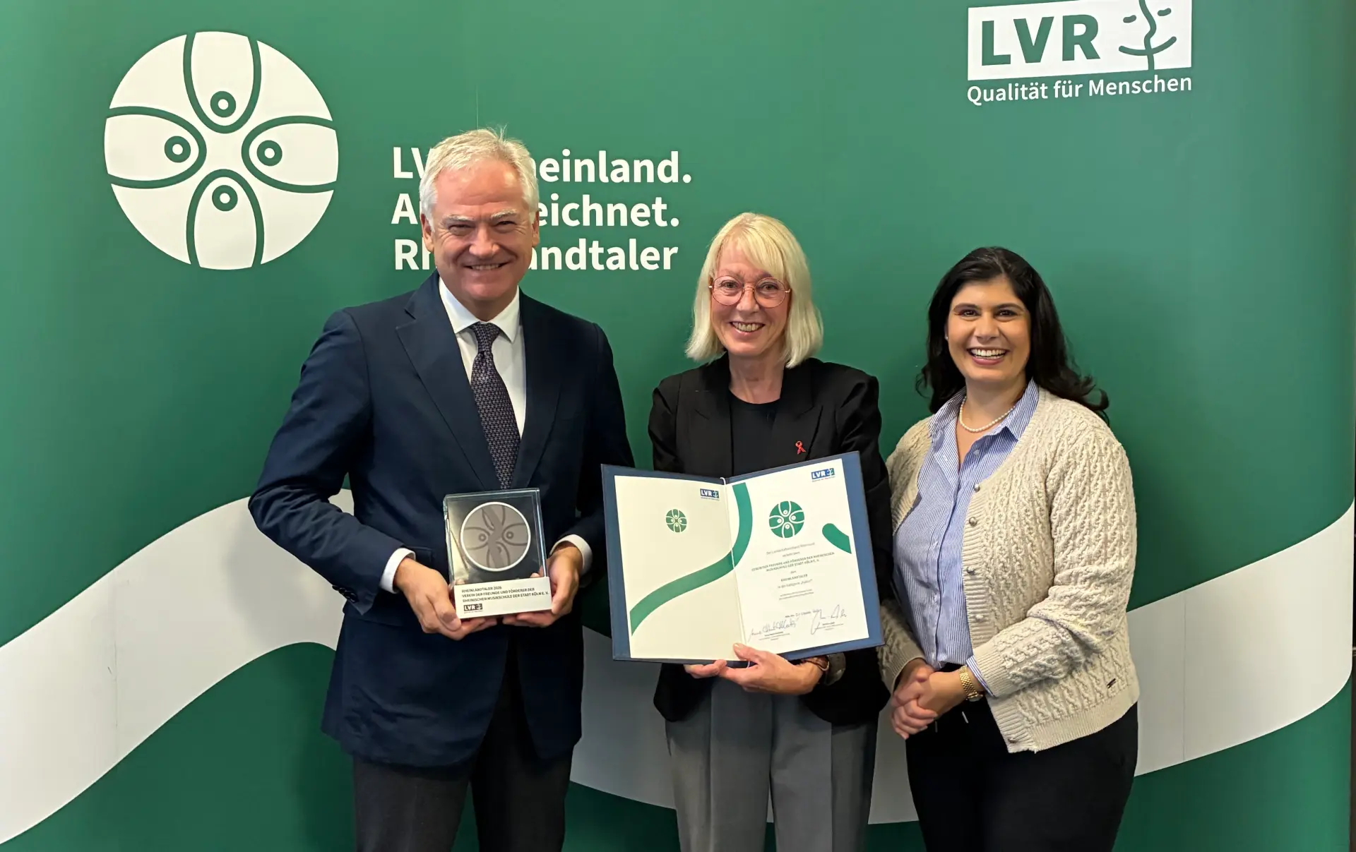 Ein Mann und zwei Frauen stehen vor einem grünen Aufsteller mit dem Rheinland ausgezeichnet-Logo