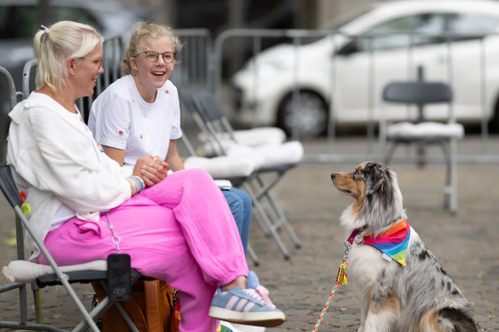Zwei Frauen sitzen auf Klappstühlen und unterhalten sich mit einem Hund, der ein Halstuch in den Farben der Progress-Pride-Flagge trägt.