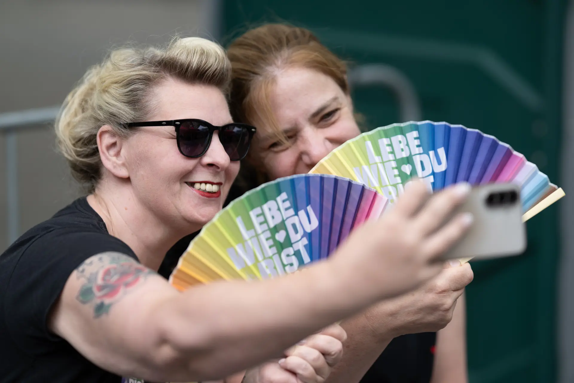 Zwei Frauen lächeln beim gemeinsamen Selfie in ein Smartphone und halten bunte Fächer mit der Aufschrift „Lebe wie du bist“ vor sich.