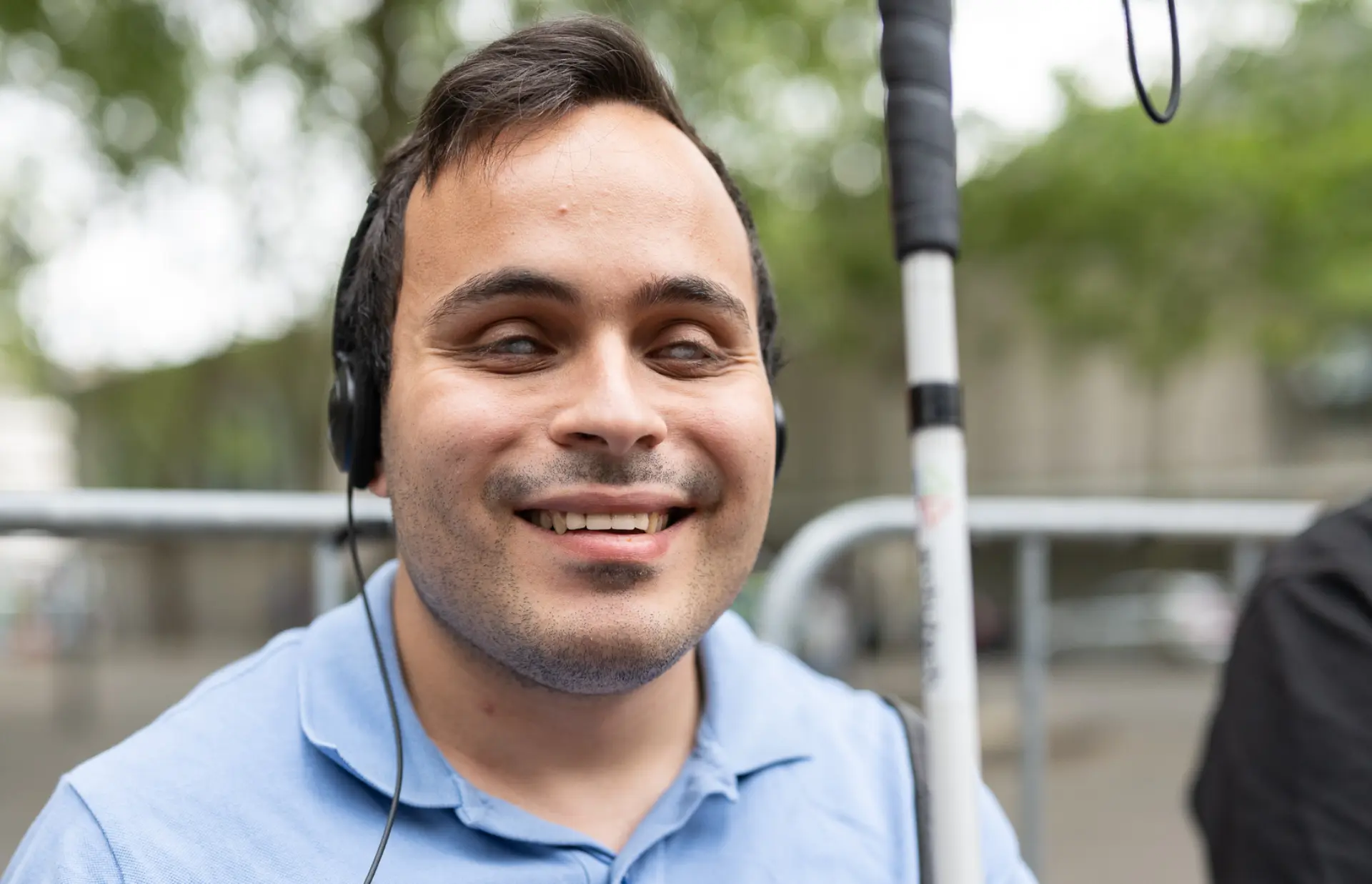 Ein blinder Mann mit Kopfhörer und Langstock lächelt in die Kamera und hält ein kleines Audiogerät in den Händen.