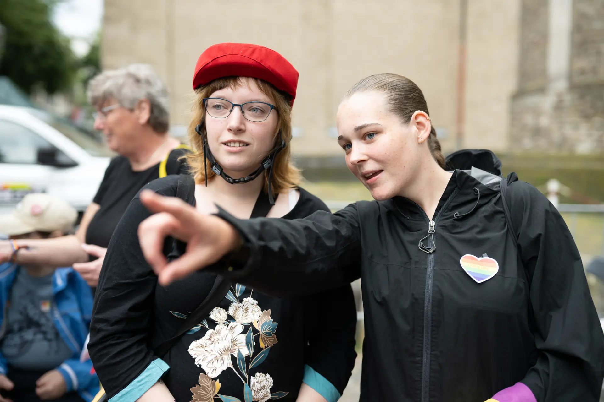 Zwei junge Frauen stehen nebeneinander; eine von ihnen trägt einen roten Schutzhelm, die andere zeigt mit ausgestrecktem Arm auf etwas außerhalb des Bildes.