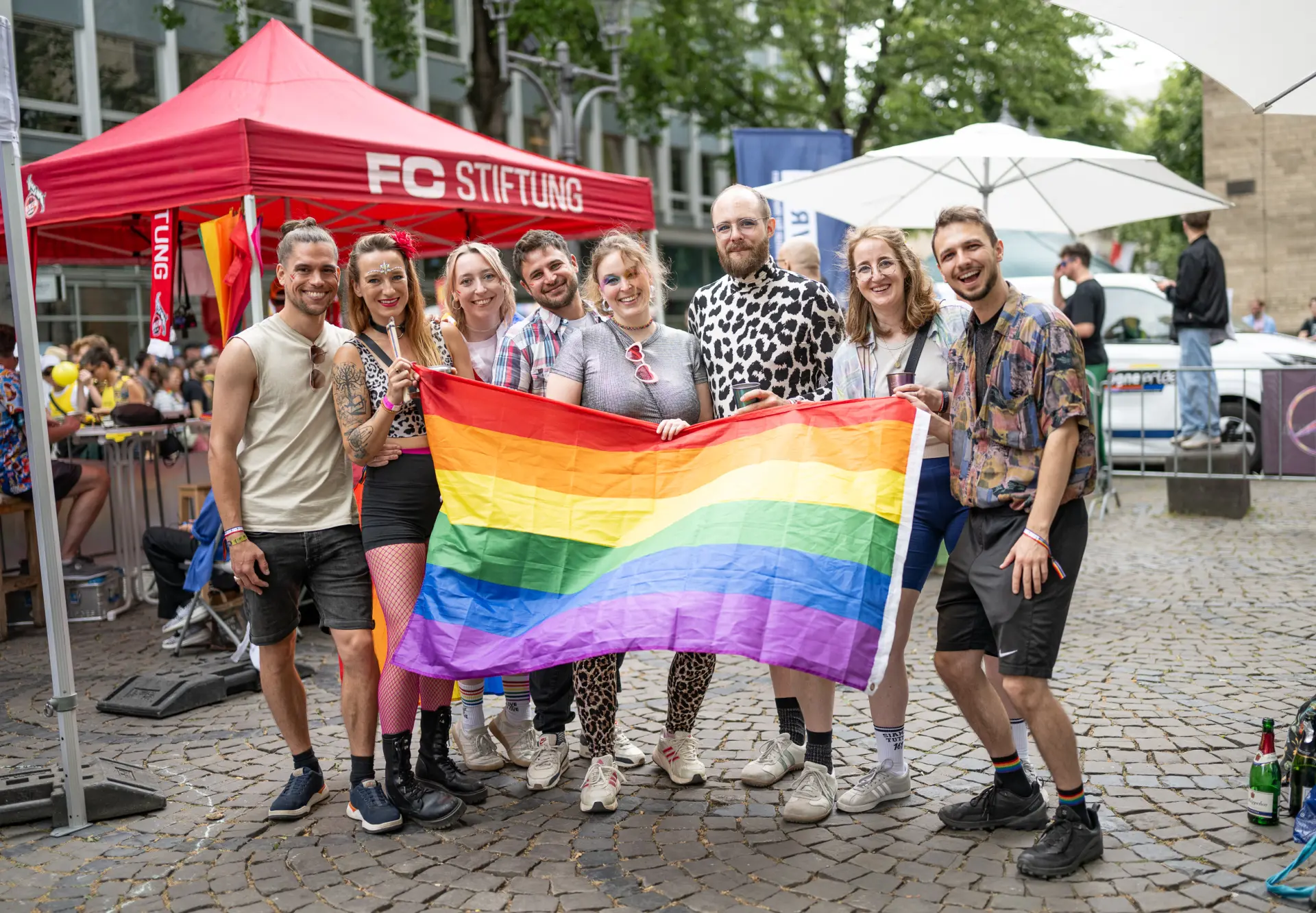 Acht Menschen stehen beim CSD nebeneinander und halten gemeinsam eine große Regenbogenflagge vor sich.