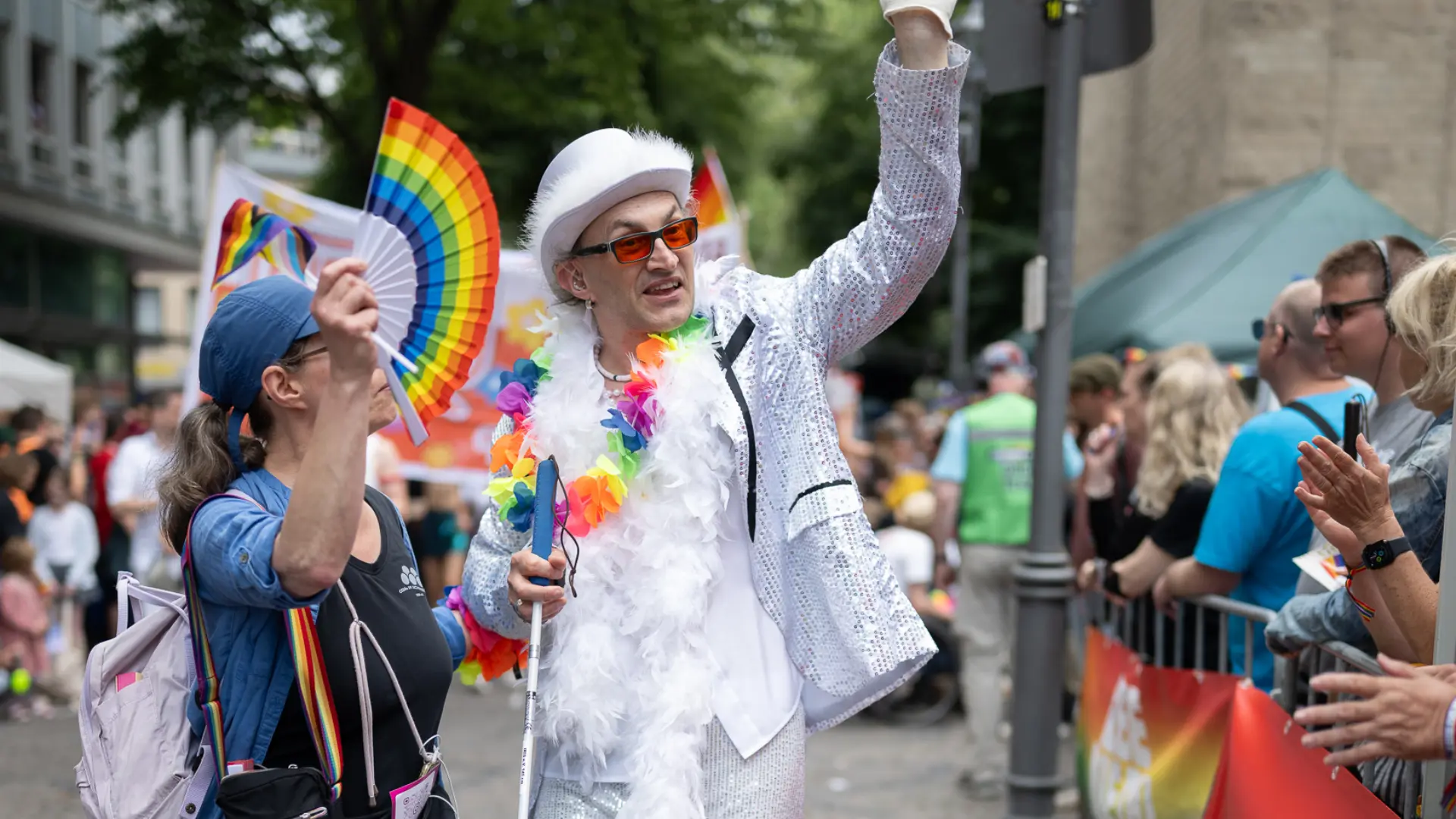 Eine Frau mit Regenbogenfächer begleitet einen weiß gekleideten Mann mit Langstock, der dem Publikum am Straßenrand zuwinkt.