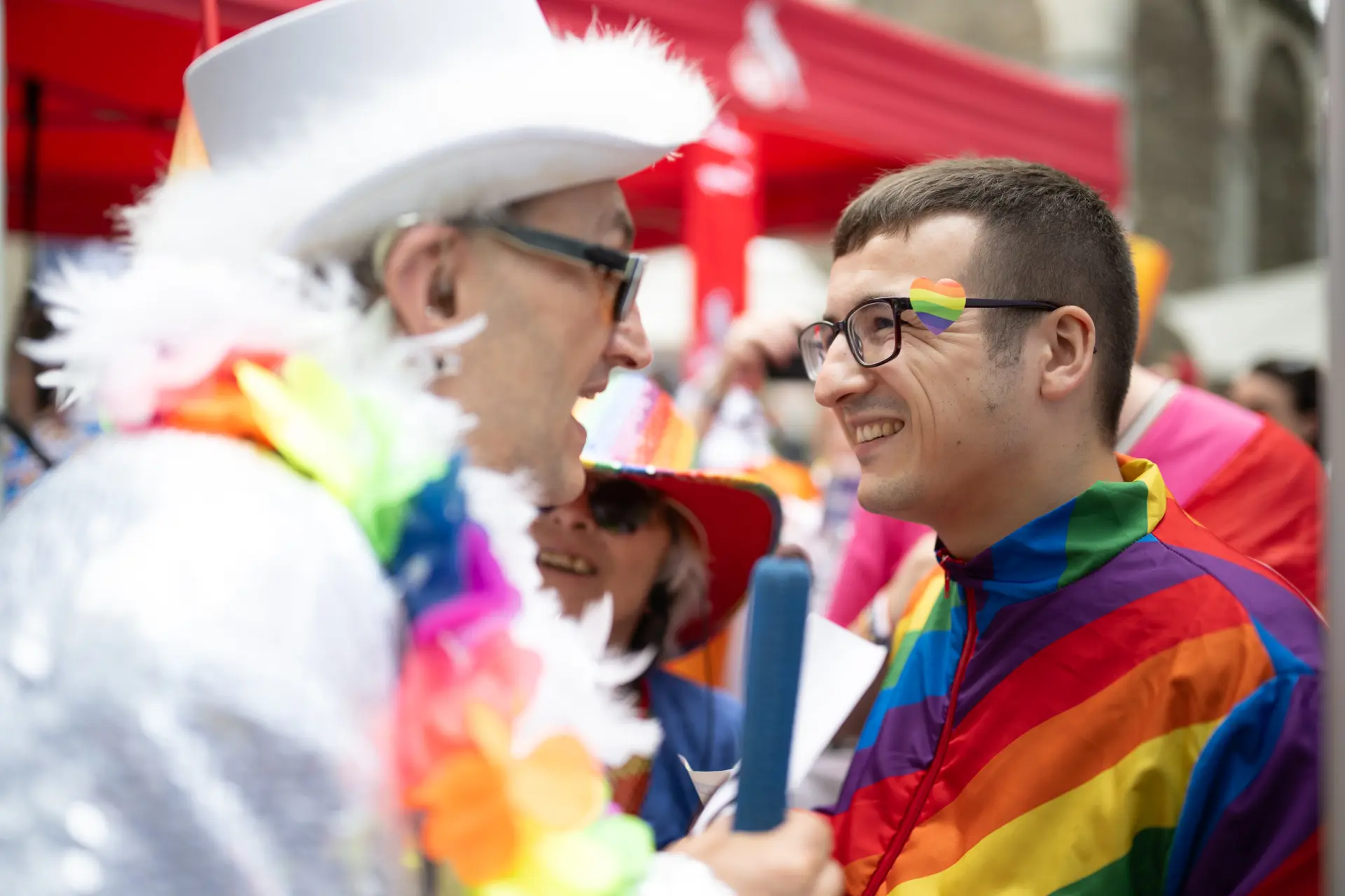 Ein Mann in einem Regenbogenanzug spricht lächelnd mit einem weiß gekleideten Mann mit Zylinder, Sonnenbrille und buntem Blumenschmuck.