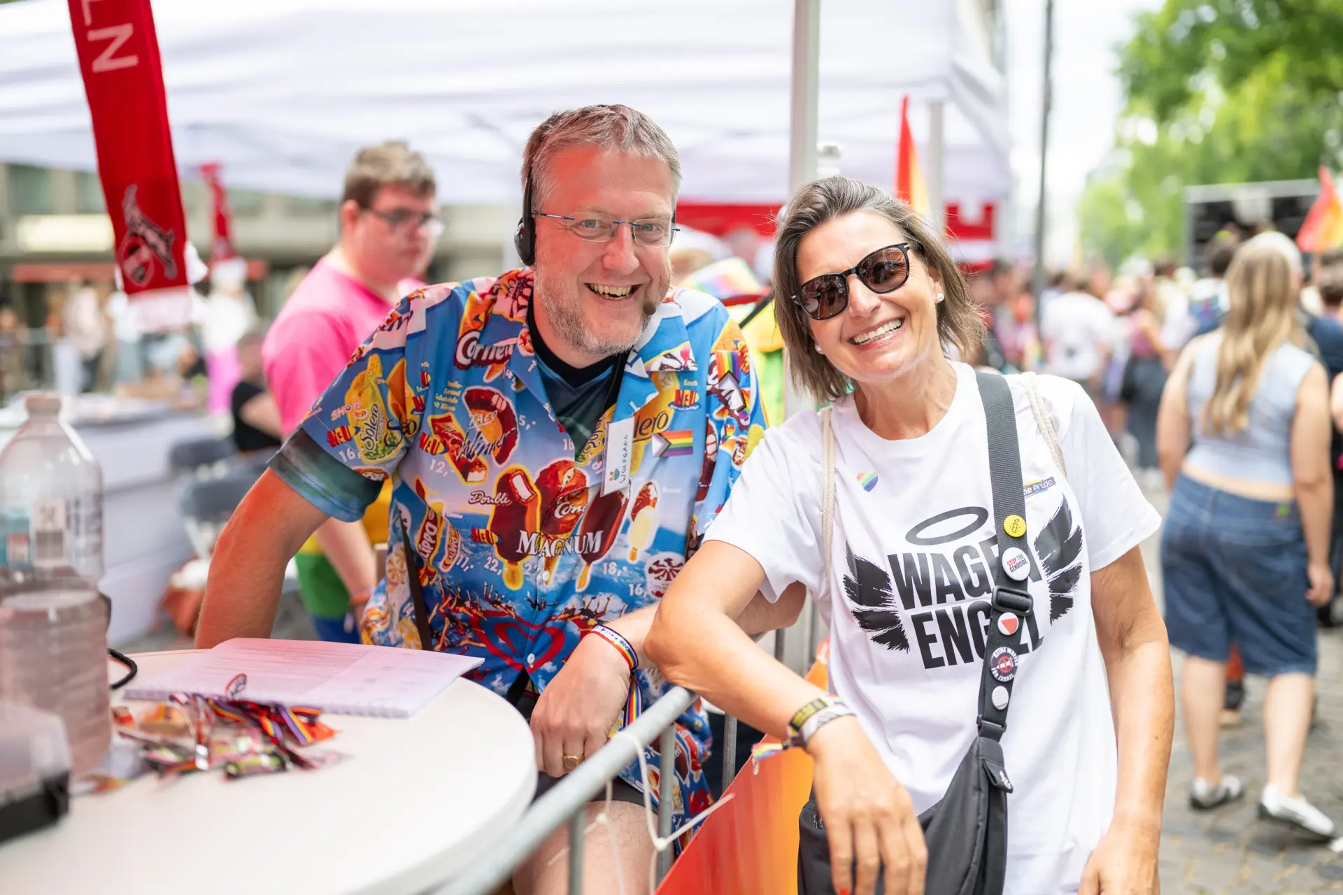 Ein Mann mit Headset und bunt bedrucktem Hemd und eine Frau mit Sonnenbrille lächeln gemeinsam an einem Stand beim CSD.