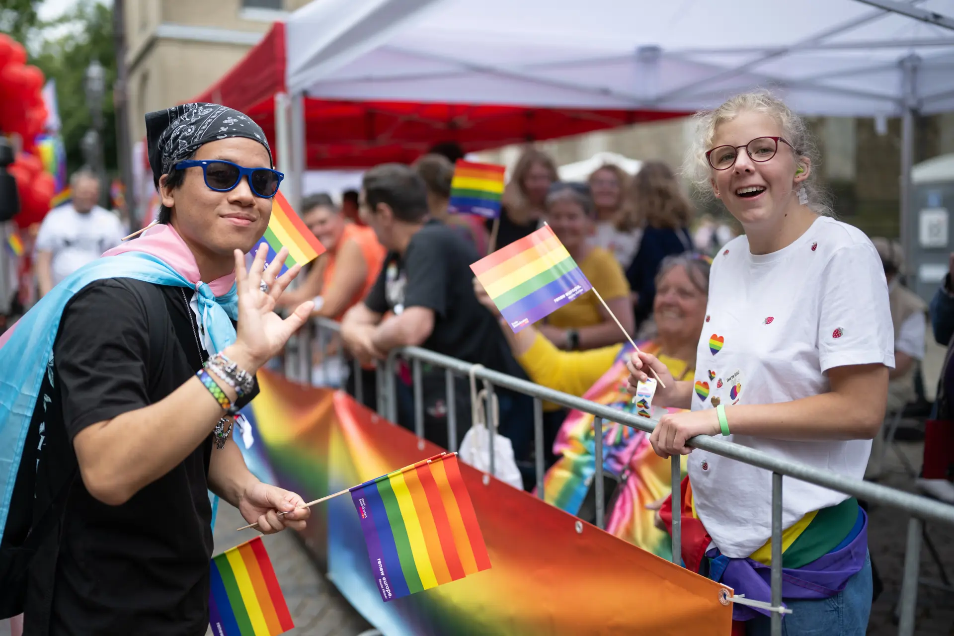 Zwei junge Menschen winken und halten Regenbogenfähnchen, während hinter ihnen weitere Teilnehmende an einem abgesperrten Veranstaltungsbereich stehen.