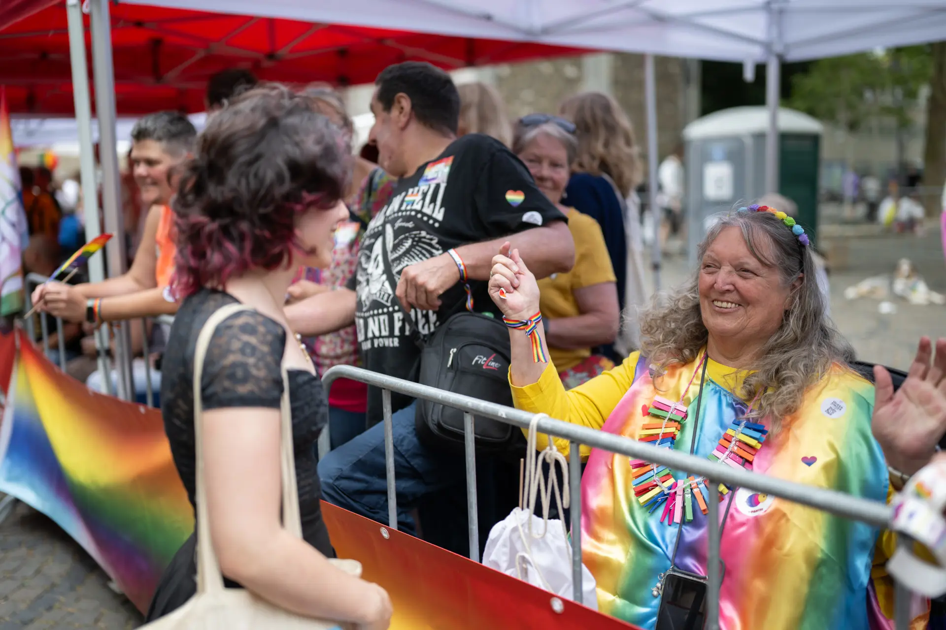 Eine ältere Frau in einem bunten Oberteil tanzt und lächelt hinter einer Absperrung mit Regenbogenbanner.