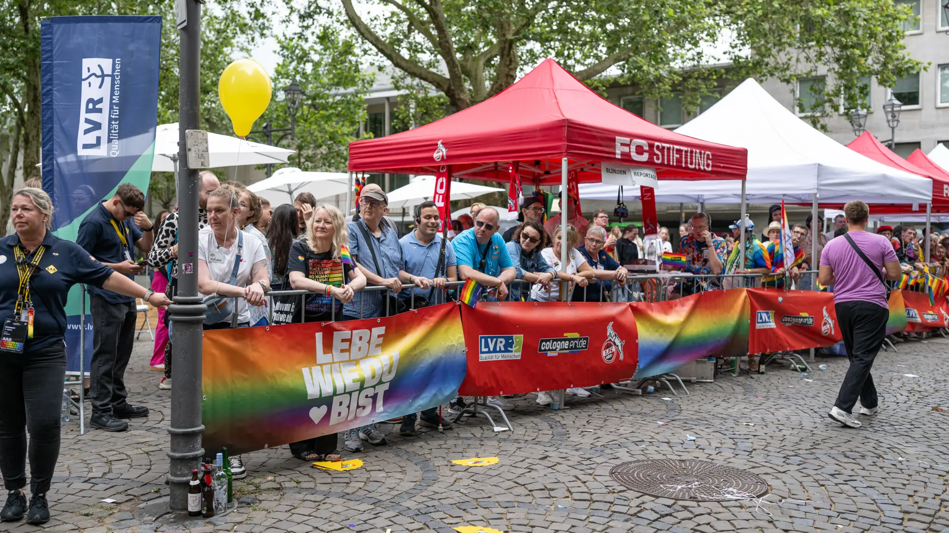 Menschen stehen beim CSD hinter einer Absperrung mit Regenbogenbanner und der Aufschrift „Lebe, wie du bist“. Im Hintergrund sind Fahnen des LVR und der FC-Stiftung zu sehen.