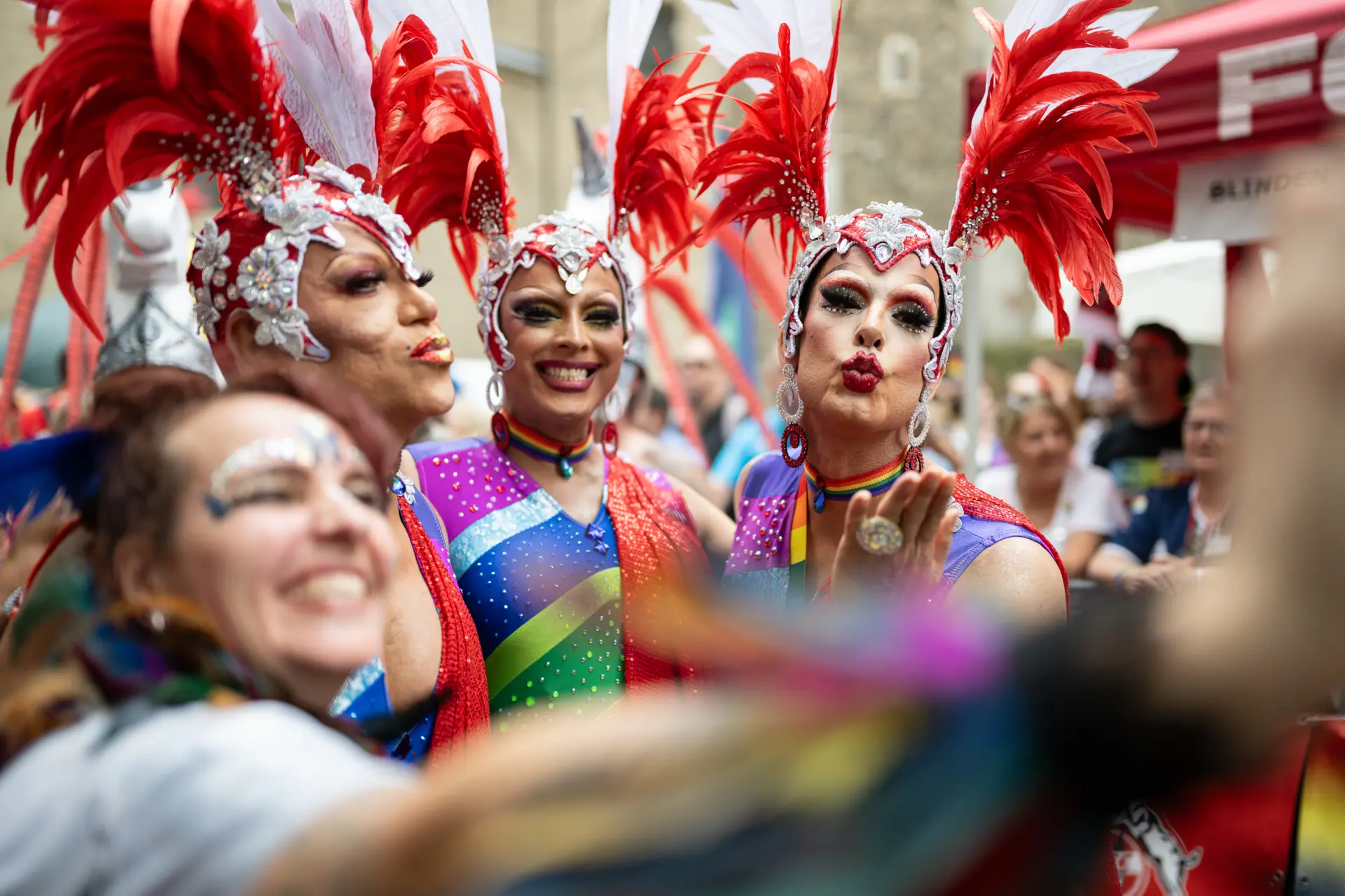 Drei Dragqueens in farbenprächtigen Kostümen mit großen rot-weißen Federkopfschmucken posieren gut gelaunt für ein gemeinsames Foto.