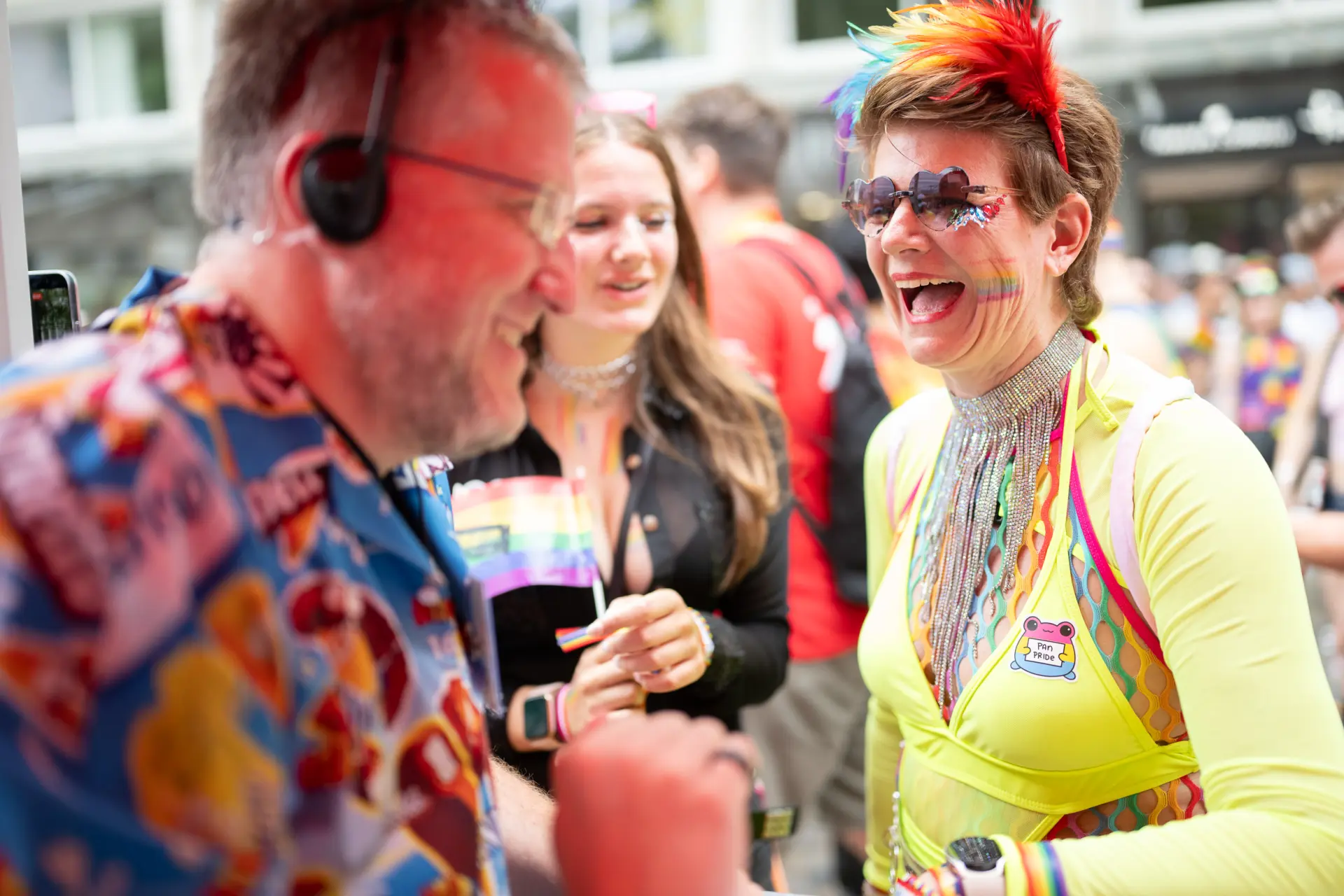 Eine bunt gekleidete Person mit herzförmiger Sonnenbrille und Regenbogenbemalung lacht im Gespräch mit zwei weiteren Menschen.