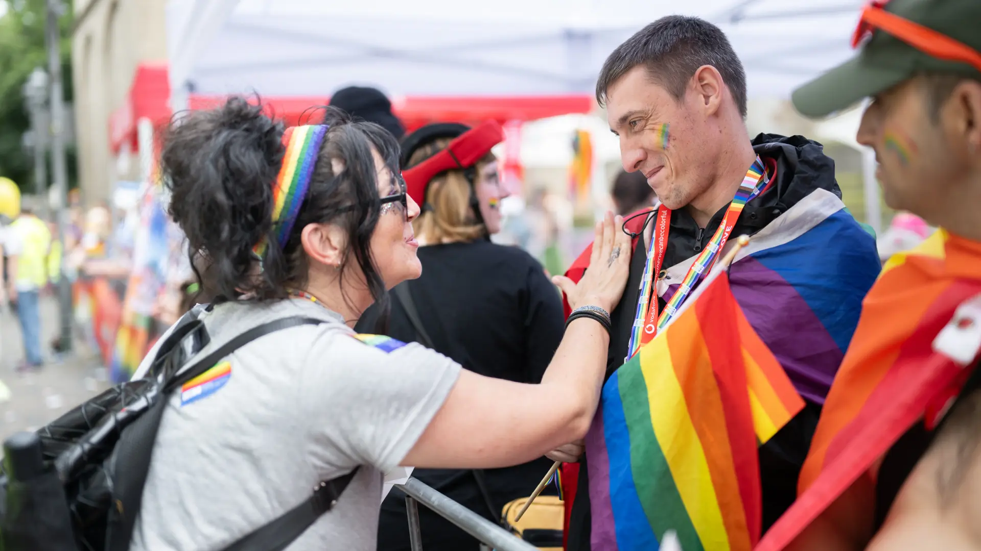 Eine Frau mit Regenbogenhaarband begrüßt einen Mann, der in eine große Regenbogenflagge gehüllt ist.