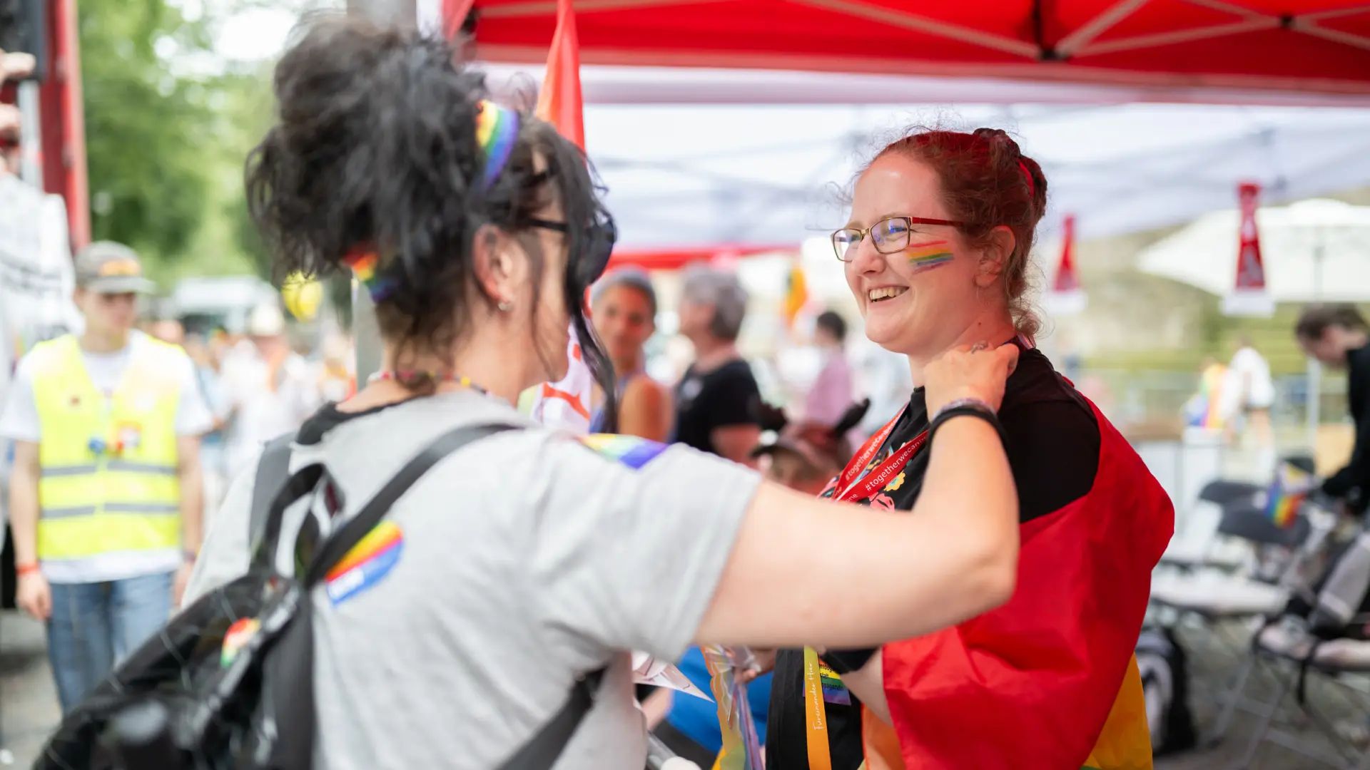 Eine Frau mit Regenbogenhaarband legt einer lachenden Frau mit roter Fahne freundschaftlich die Hände auf die Schultern.