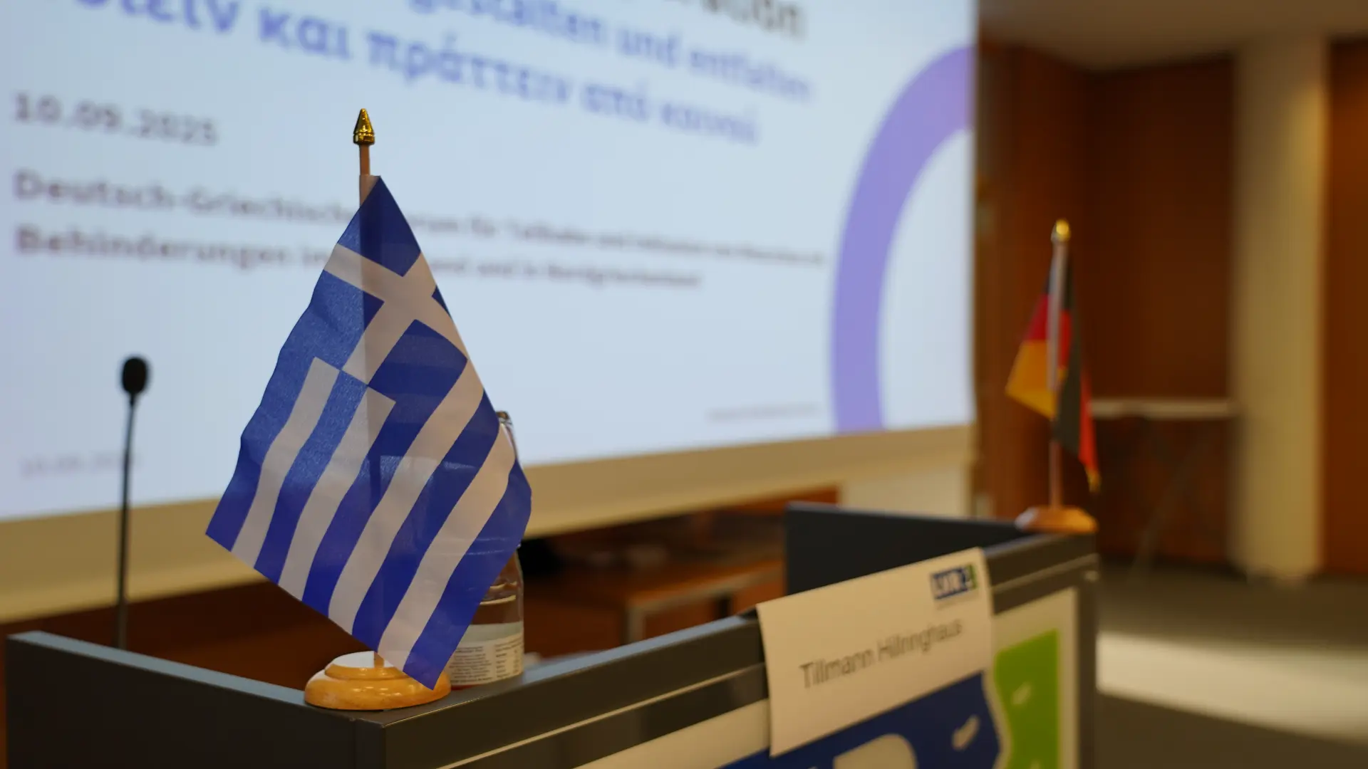 Eine griechische und eine deutsche Flagge stehen auf einem Redepult. Im Hintergrund ist eine Leinwand.