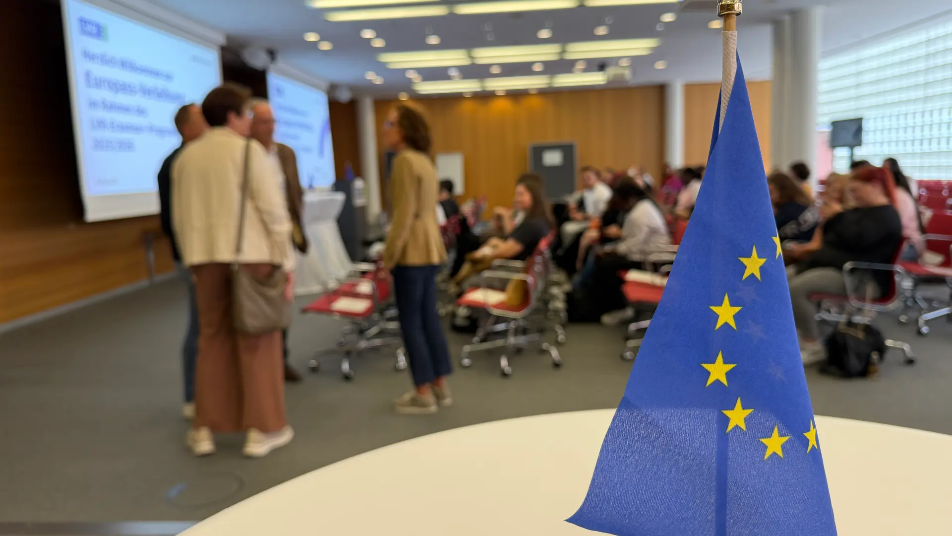 Im Vordergrund steht eine EU-Flagge auf einem Tisch. Im Hintergrund sieht man verschwommen Menschen zusammenstehen.
