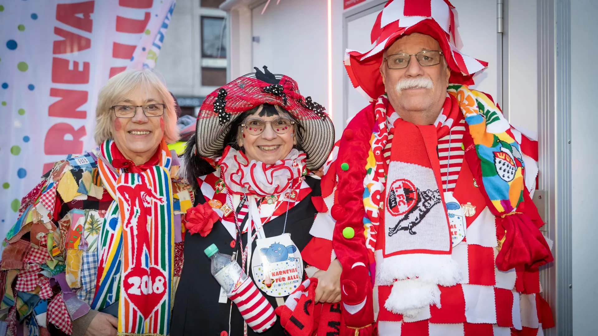 Drei Gäste beim Rosenmontag auf der LVR-Tribüne. Sie tragen bunte Kostüme und schauen fröhlich in die Kamera.