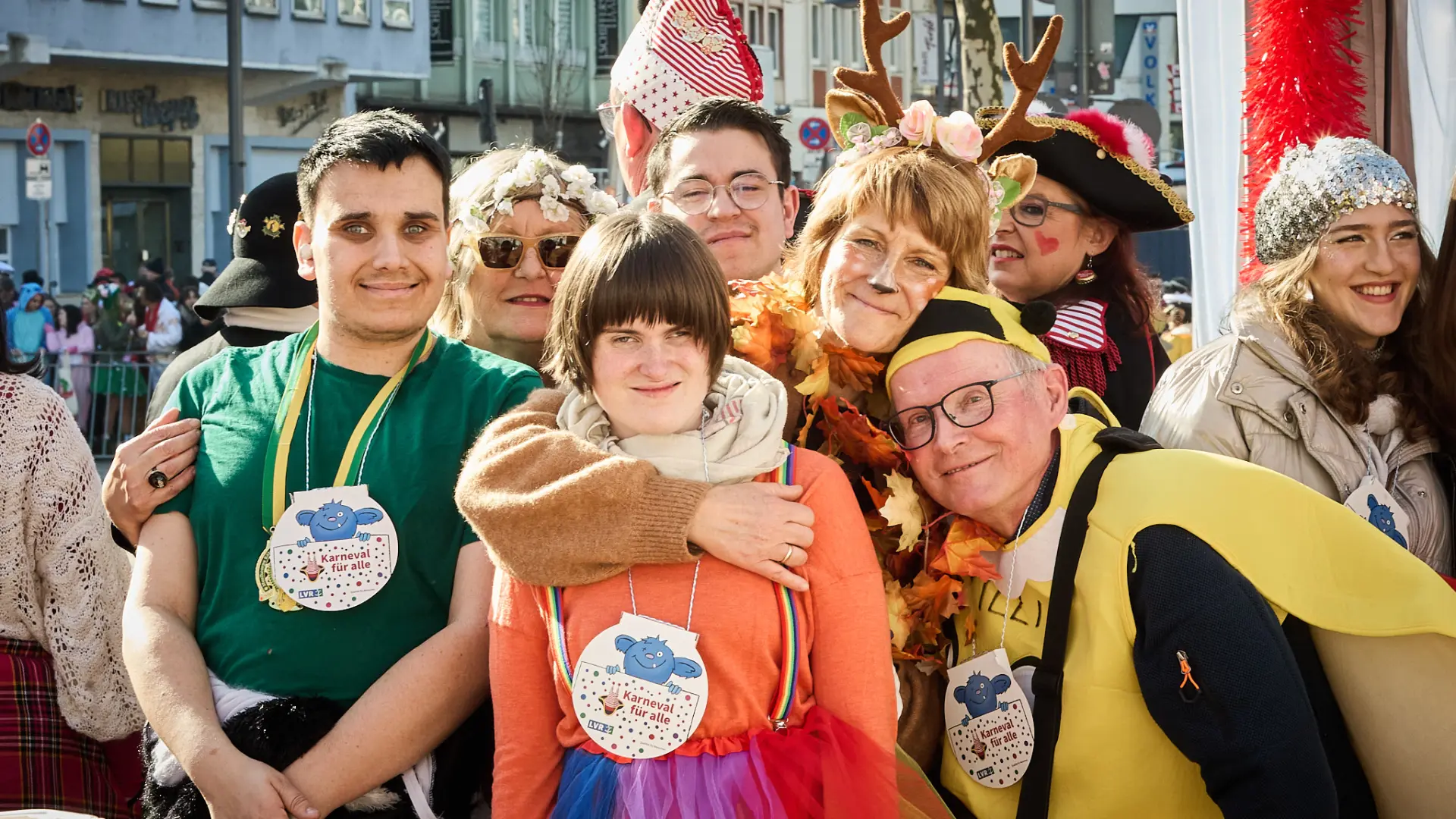 Eine Gruppe von verkleideten jungen Menschen steht vor der LVR-Tribüne bei den Schull- und Veedelszöch in Köln. Sie tragen einen Orden der Initiative "Karneval für alle".