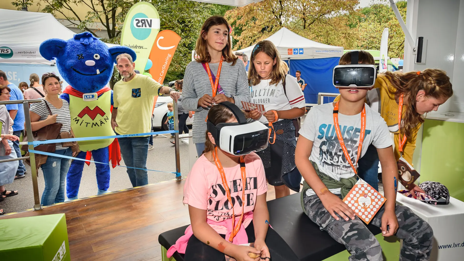 Kinder testen auf dem "Mobil der Begegnung" die VR-Brillen. Im Hintergrund ist Mitmän zu sehen, das lebensgroße LVR-Maskottchen aus blauem Plüsch. 