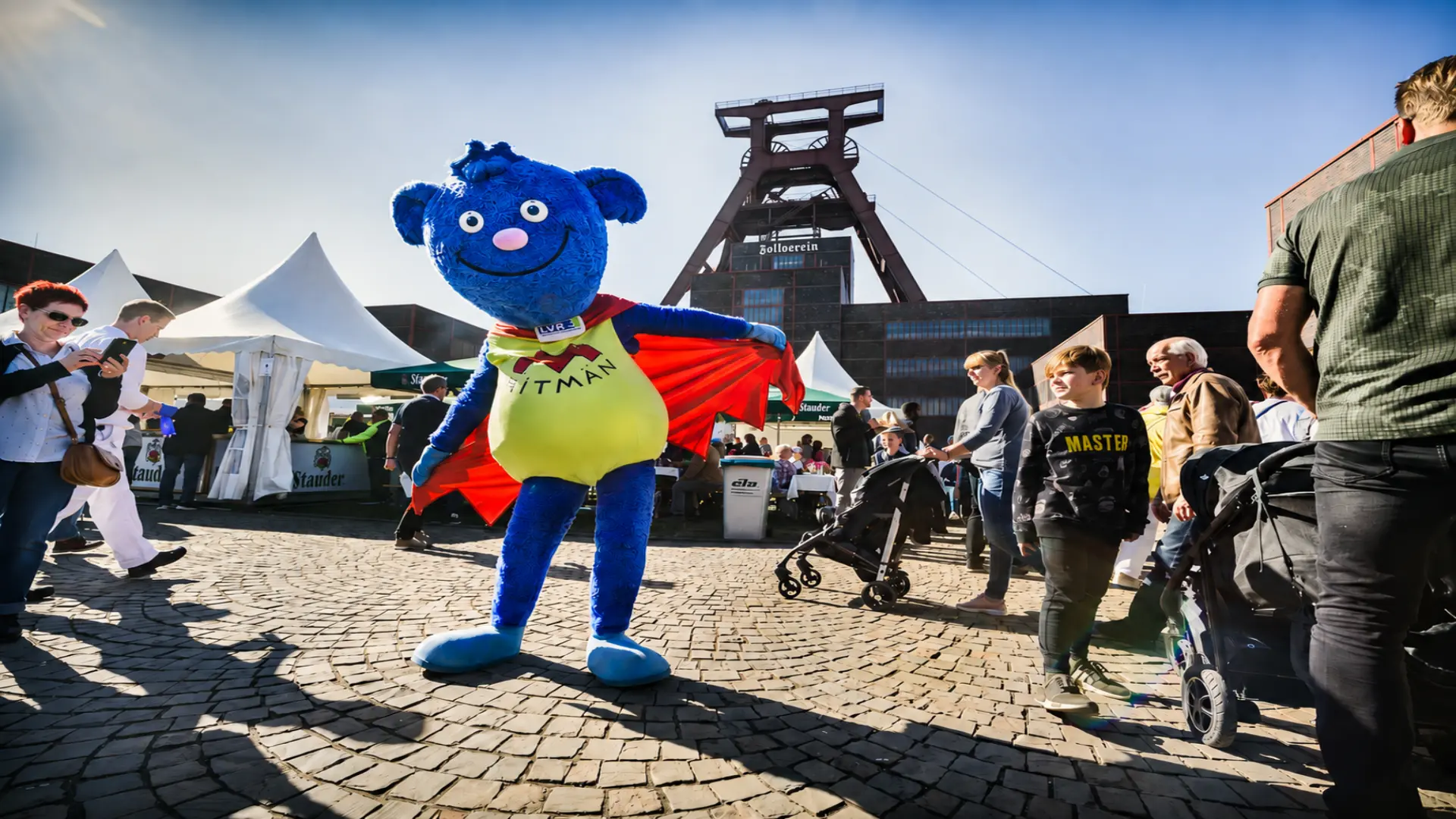 Das Foto zeigt das LVR-Maskottchen Mitmän. Es tanzt vor der Zeche Zollverein. 