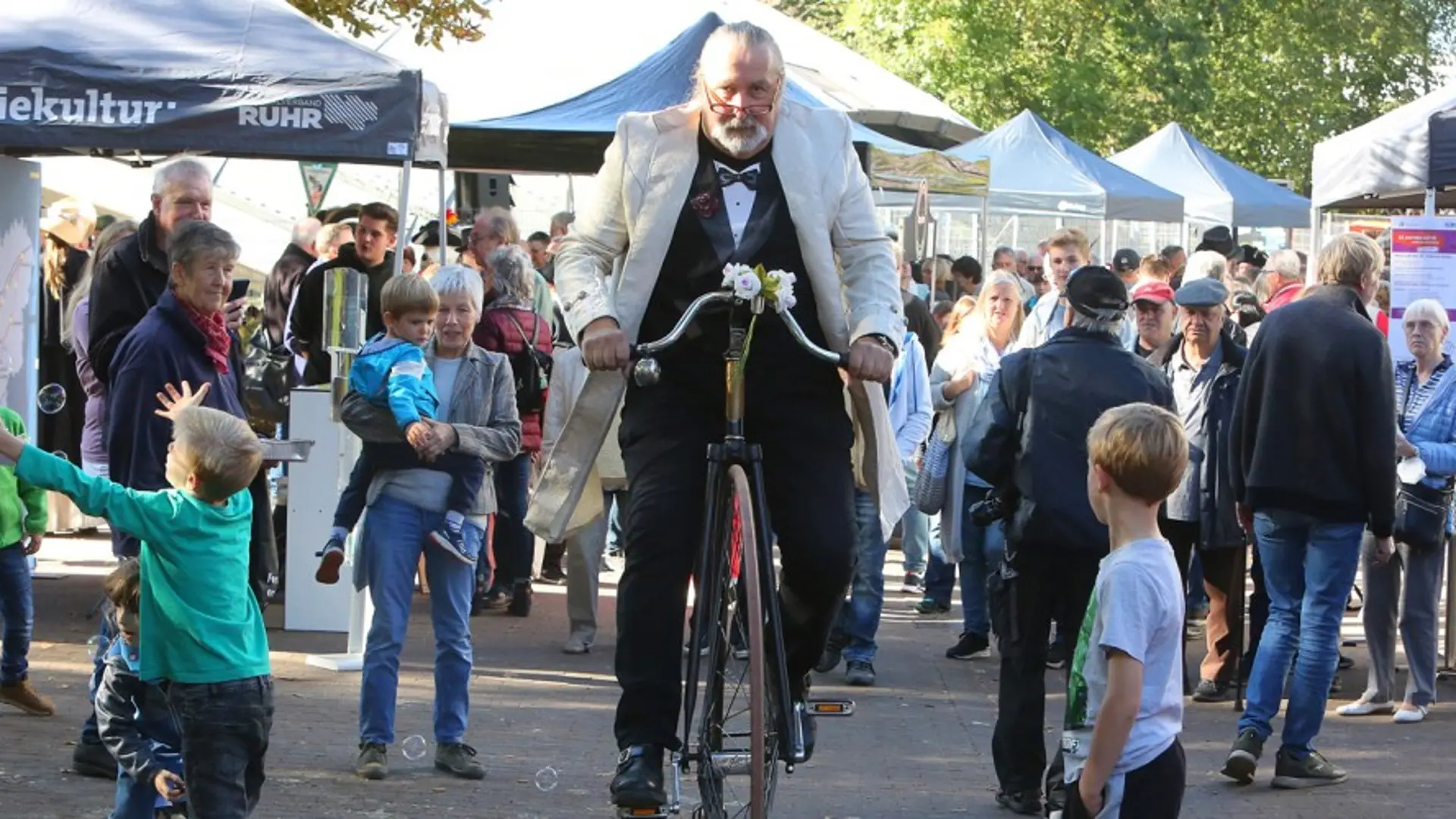 Das Bild zeigt einen historischen Jahrmarkt. Im Mittelpunkt ist ein Mann zu sehen, der auf einem Hochrad aus dem 19. Jahrhundert  sitzt. 