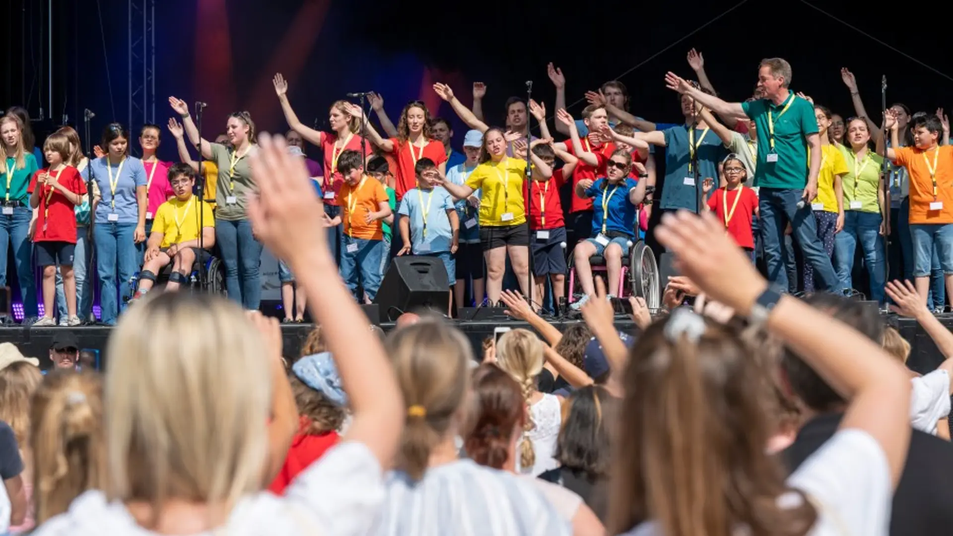 Das Foto zeigt einen großen Kinderchor in bunter Kleidung auf einer großen Bühne.