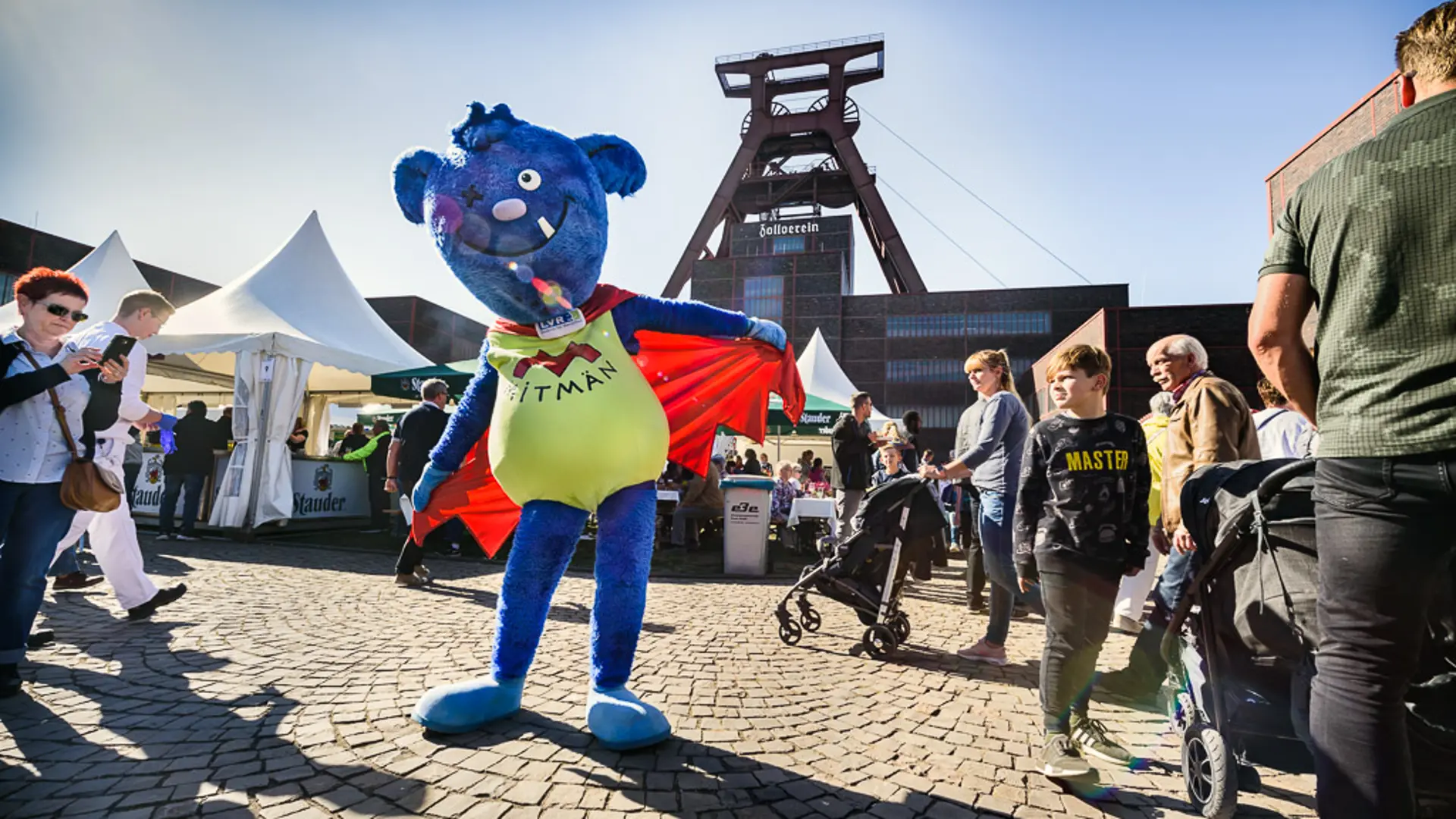 Das Foto zeigt das LVR-Maskottchen Mitmän. Es tanzt vor der Zeche Zollverein. 