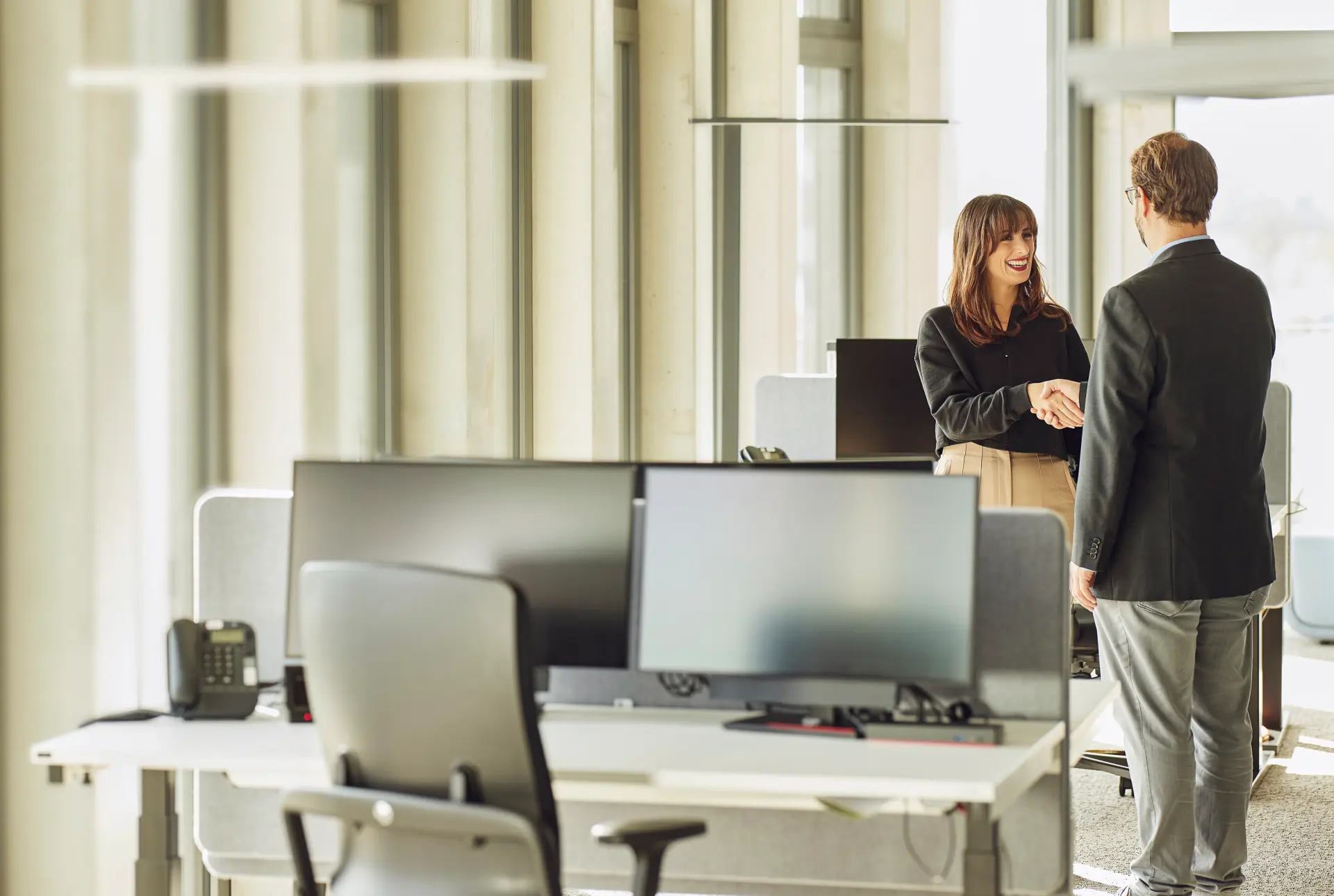 Zwei Menschen stehen sich in einem hellen Büro gegenüber und geben sich zur Begrüßung die Hand. Eine Frau lächelt, während beide Businesskleidung tragen. Im Hintergrund sind Fenster und ein Arbeitsplatz zu sehen.
