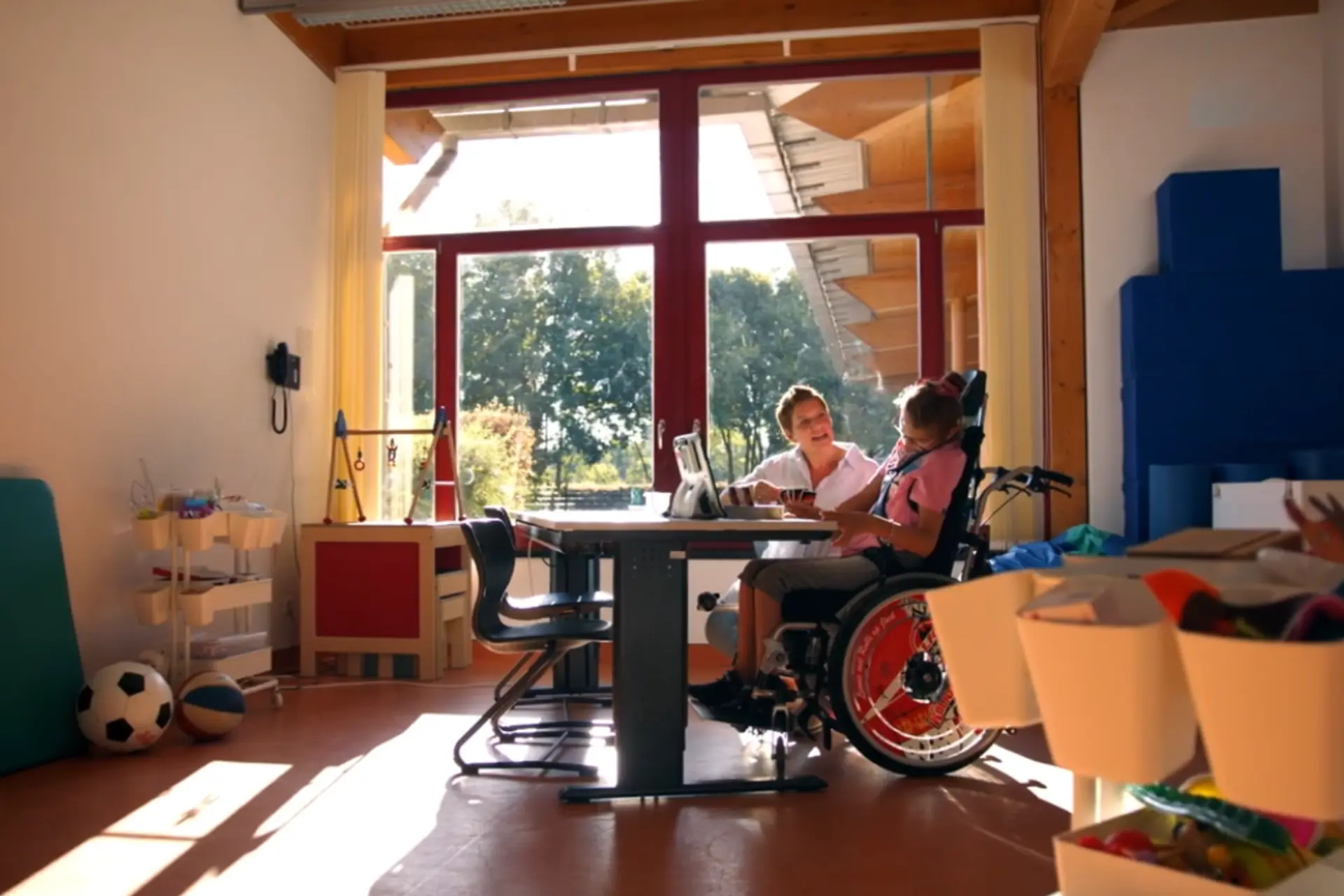 Eine Frau in einem weißen Kittel und ein Mädchen im Rollstuhl sitzen gemeinsam in einem lichtdurchfluteten Raum an einem Tisch und spielen Karrten. 