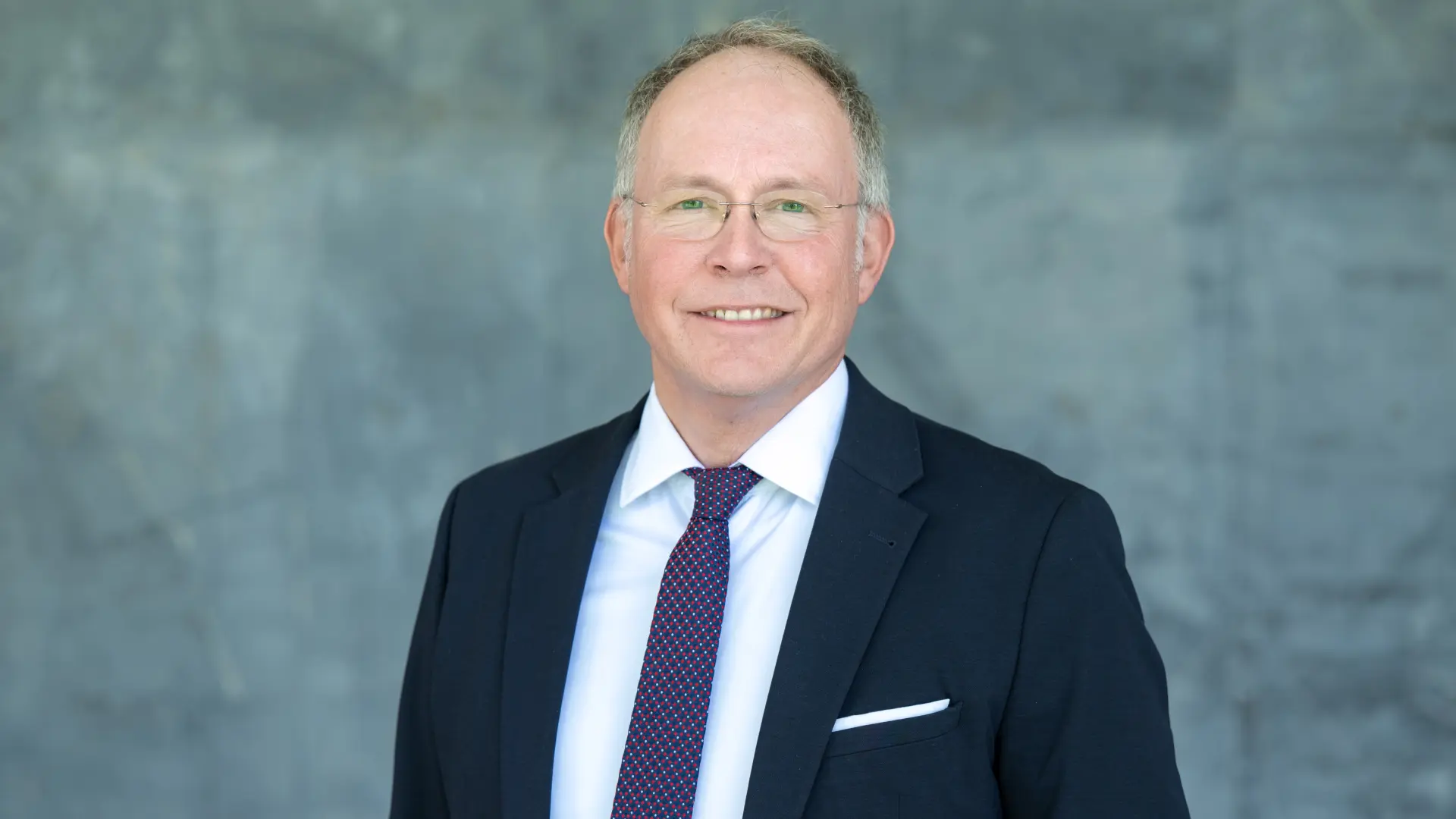 Lächelnder Mann in dunklem Anzug vor einer grauen Marmorwand.