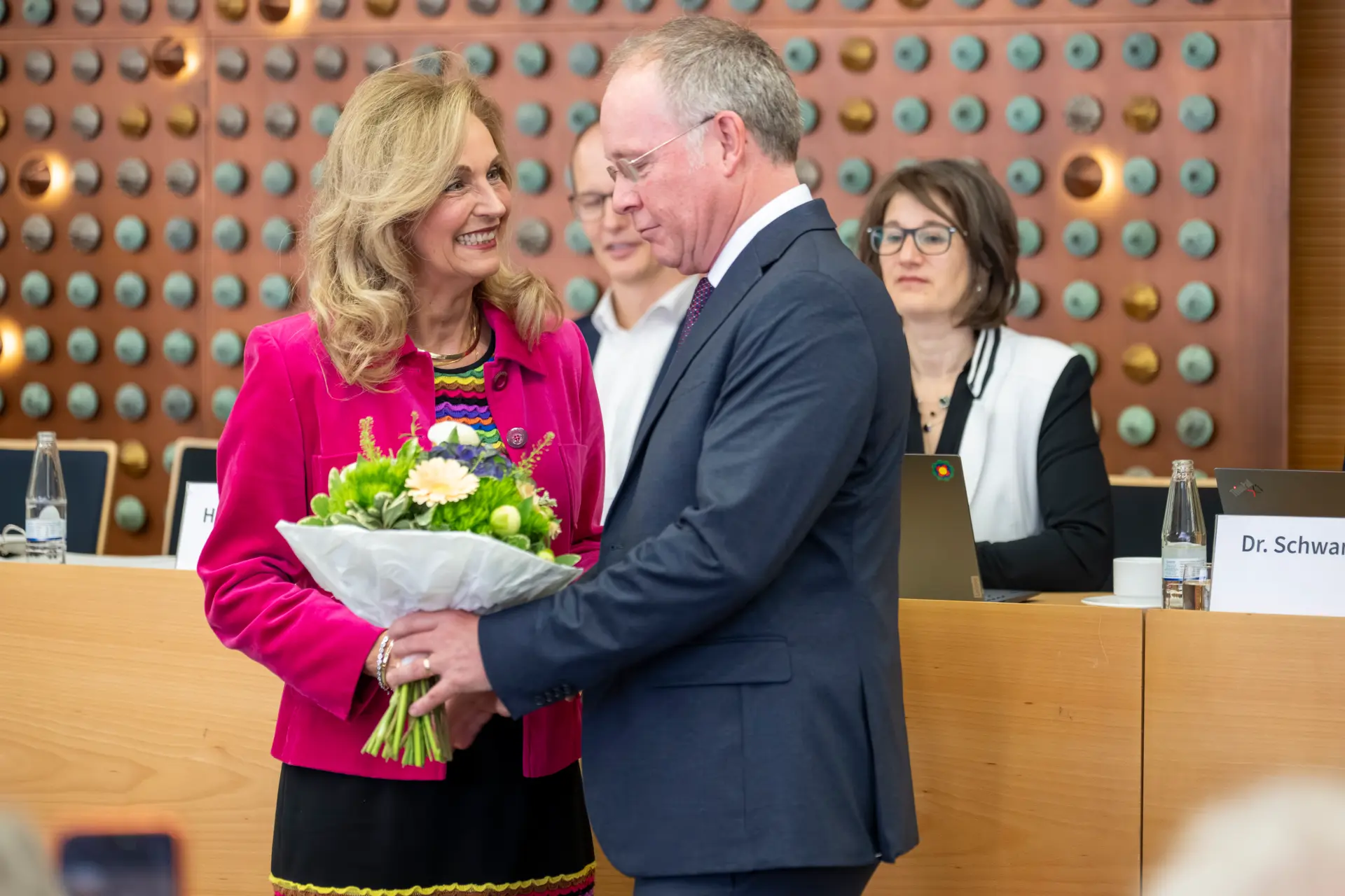 Eine lächelnde blonde Frau in pinkfarbener Jacke hält einen Blumenstrauß mit weißen, gelben und violetten Blüten. Ein Mann im dunkelblauen Anzug steht ihr gegenüber und überreicht oder richtet den Strauß. Im Hintergrund sitzen zwei Personen an einem Veranstaltungstisch mit Laptops, Wasserflaschen und Namensschildern. Die Wand dahinter ist mit vielen runden Elementen in Türkis, Grau und Gold gestaltet.