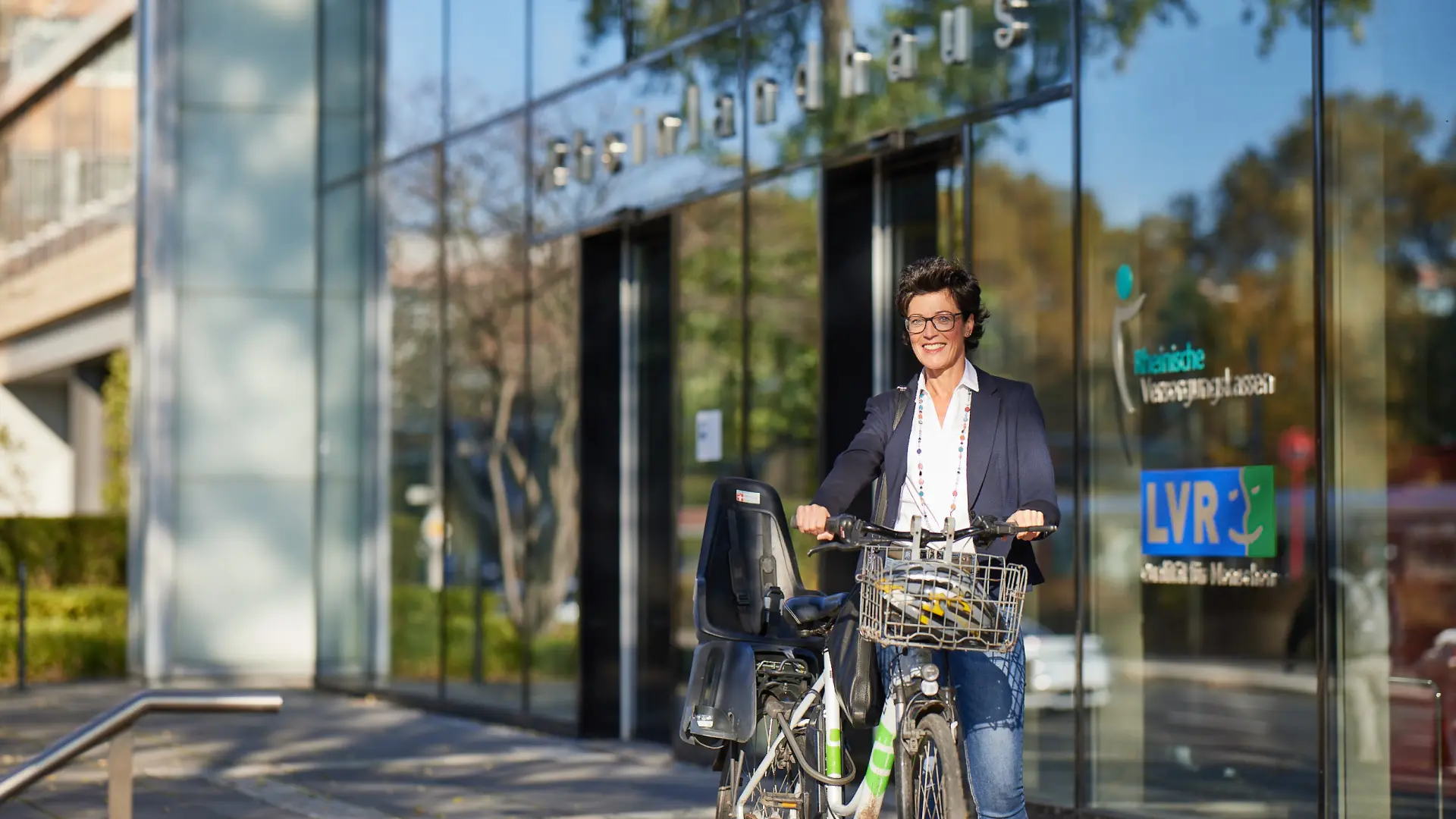 Eine lächelnde Frau schiebt ein Fahrrad. Hinter ihr steht das LVR-Rheinlandhaus.