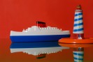 Ein Miniatur-Dampfer in blau und weiß neben einem gestreiften Miniatur-Leuchtturm vor rotem Hintergrund.